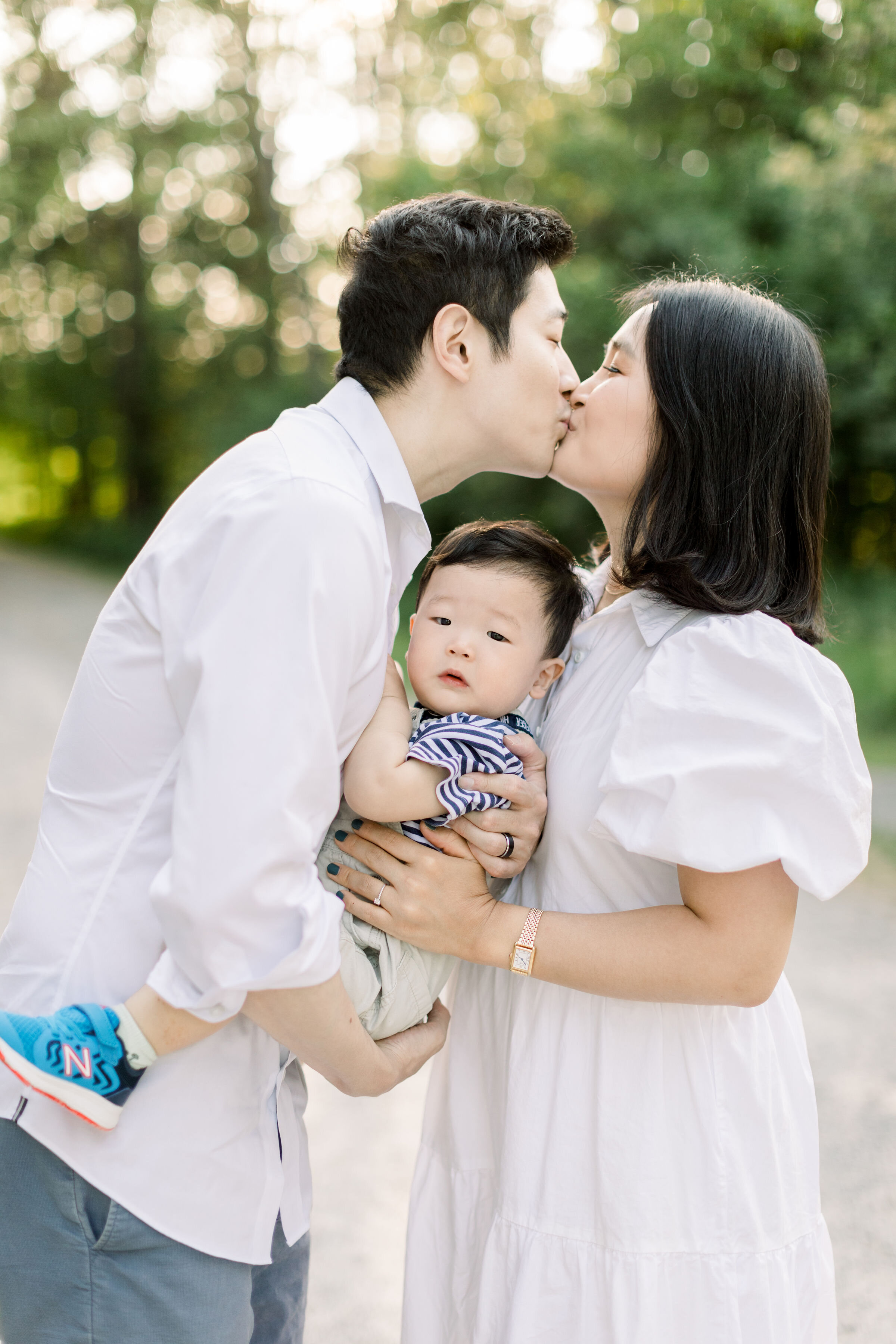  Ottawa, Canada family photographer, Chelsea Mason photography this couple holding their toddler son between them as they lean in for a kiss. kissing couple with toddler boy Ottawa family photographer white family photo outfits women's white tiered d