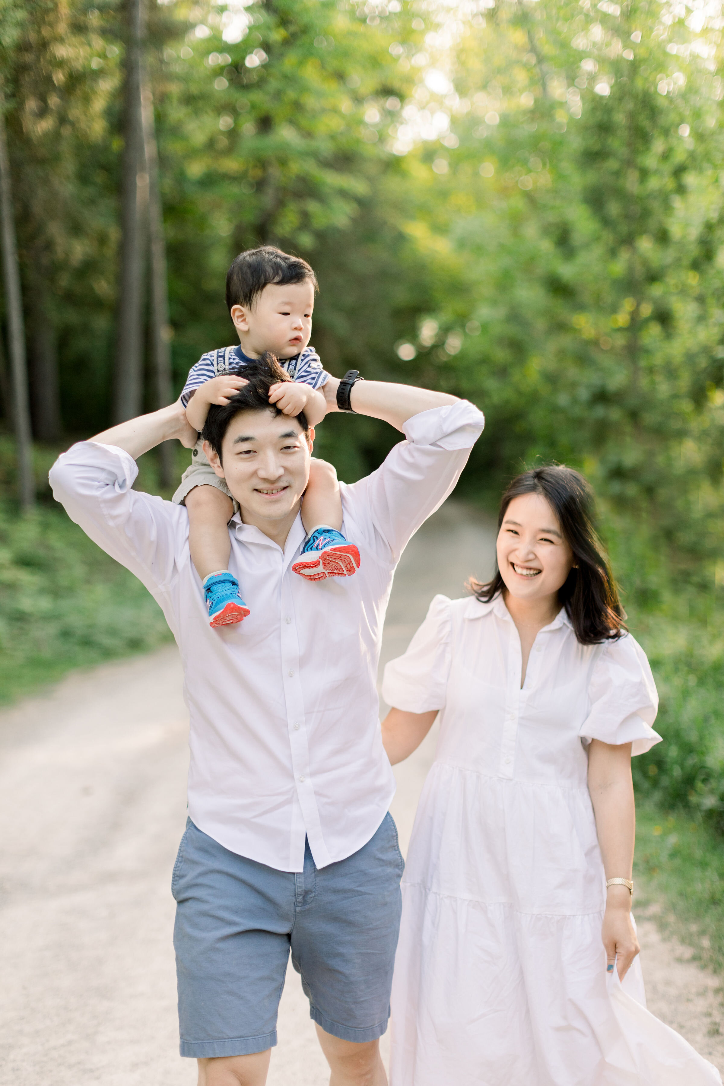  While walking down a shaded gravel pathway, Ottawa, Ontario photographer, Chelsea Mason Photography captures this father playfully giving his toddler son a piggy back ride while the mother laughs beside them. laughing family photos Ottawa Ontario fa