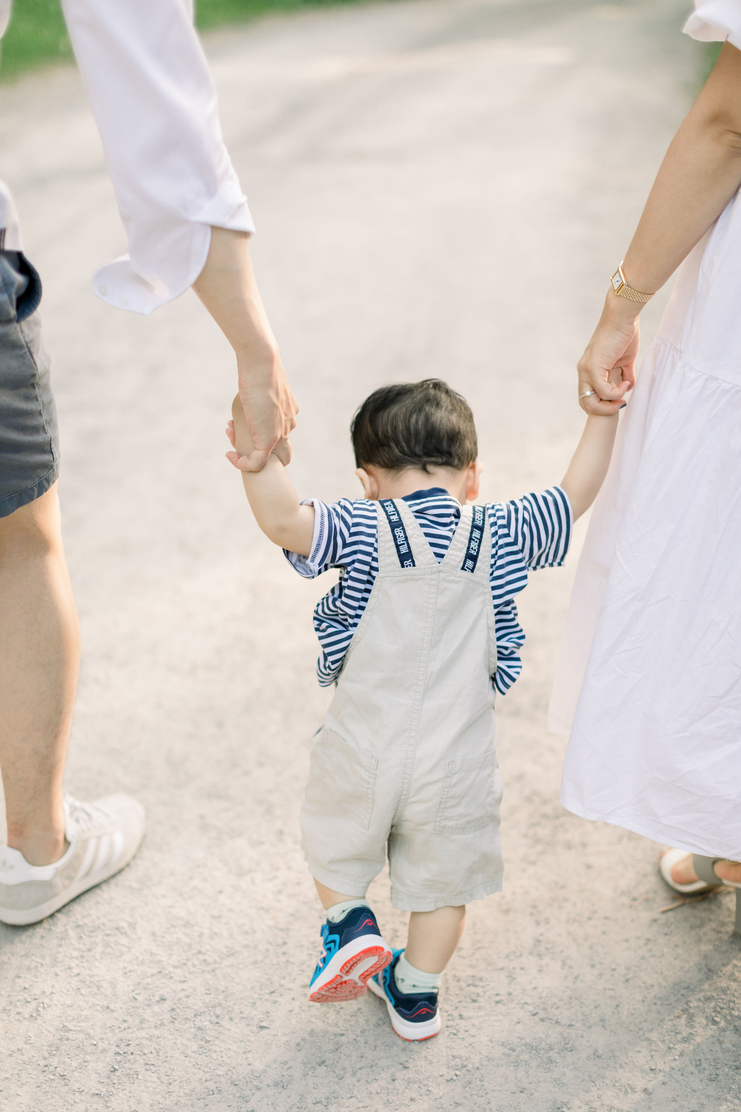  While holding both his parents hands and walking down a gravel shaded pathway, Ottawa, Ontario's Chelsea Mason Photography captures this toddler boy walking. toddler boy walking while holding parents hands toddler boy overalls with striped shirt neu