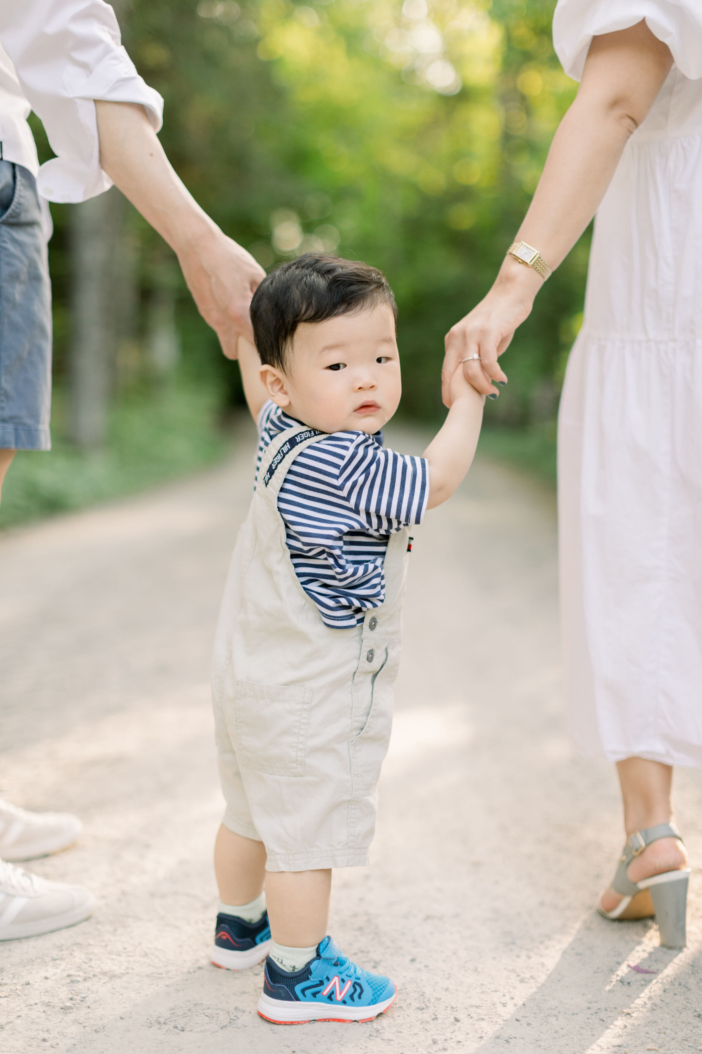  While holding both his parents hands and walking down a gravel shaded pathway, Ottawa, Ontario's Chelsea Mason Photography captures an up close candid photo. up-close toddler portrait holding parents hands Ottawa photographer #AlmonteOntarioFamilyPh