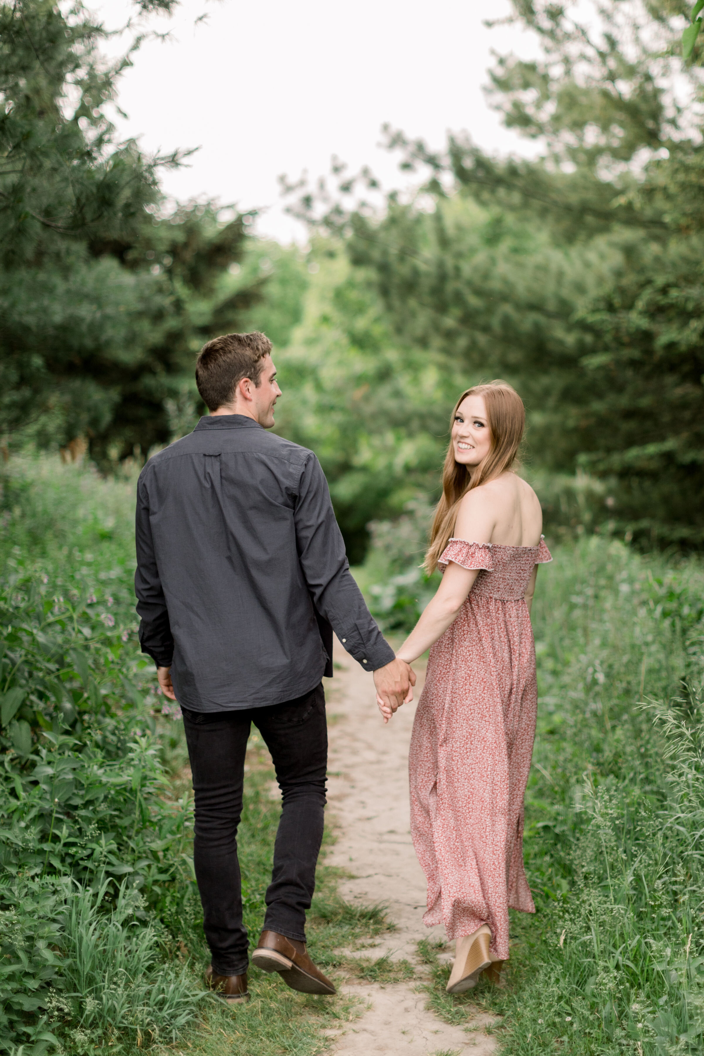  Arboretum, Ottawa photographer, engagement photographer, Chelsea Mason Photography captures this couple from behind while walking down an unpaved clearing together with their arms wrapped around one another. woman looking over her shoulder smiling c