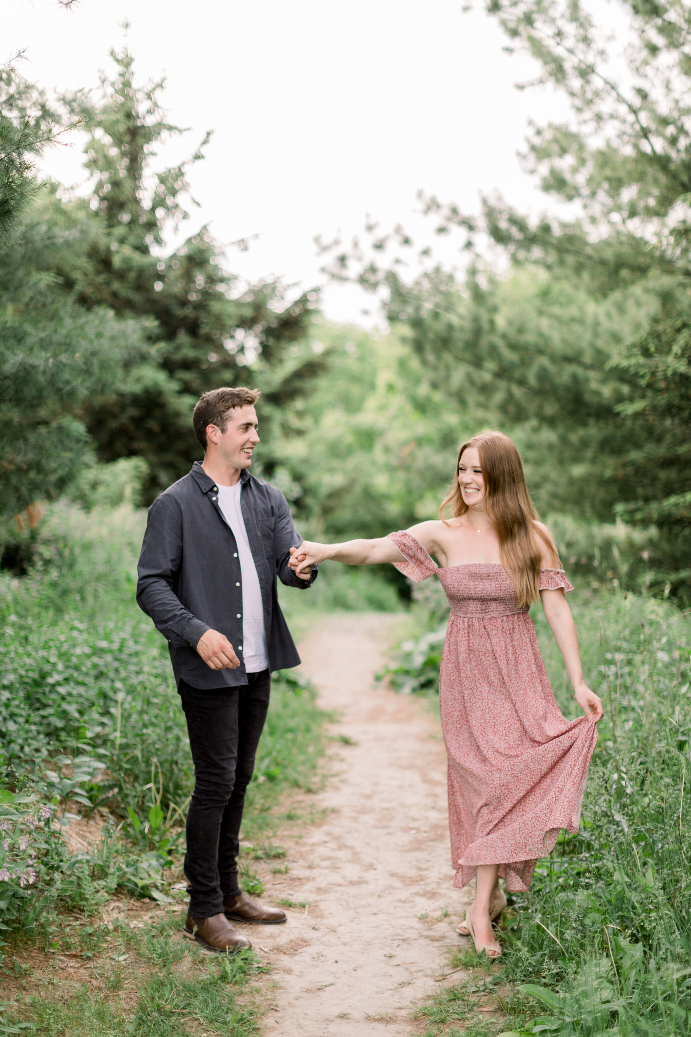  In Arboretum, Ottawa, Chelsea Mason Photography captures this couple playfully holding hands and walking down a shaded unpaved pathway. women's off the shoulder dress formal engagement outfits couple holding hands arboretum photographer women's long