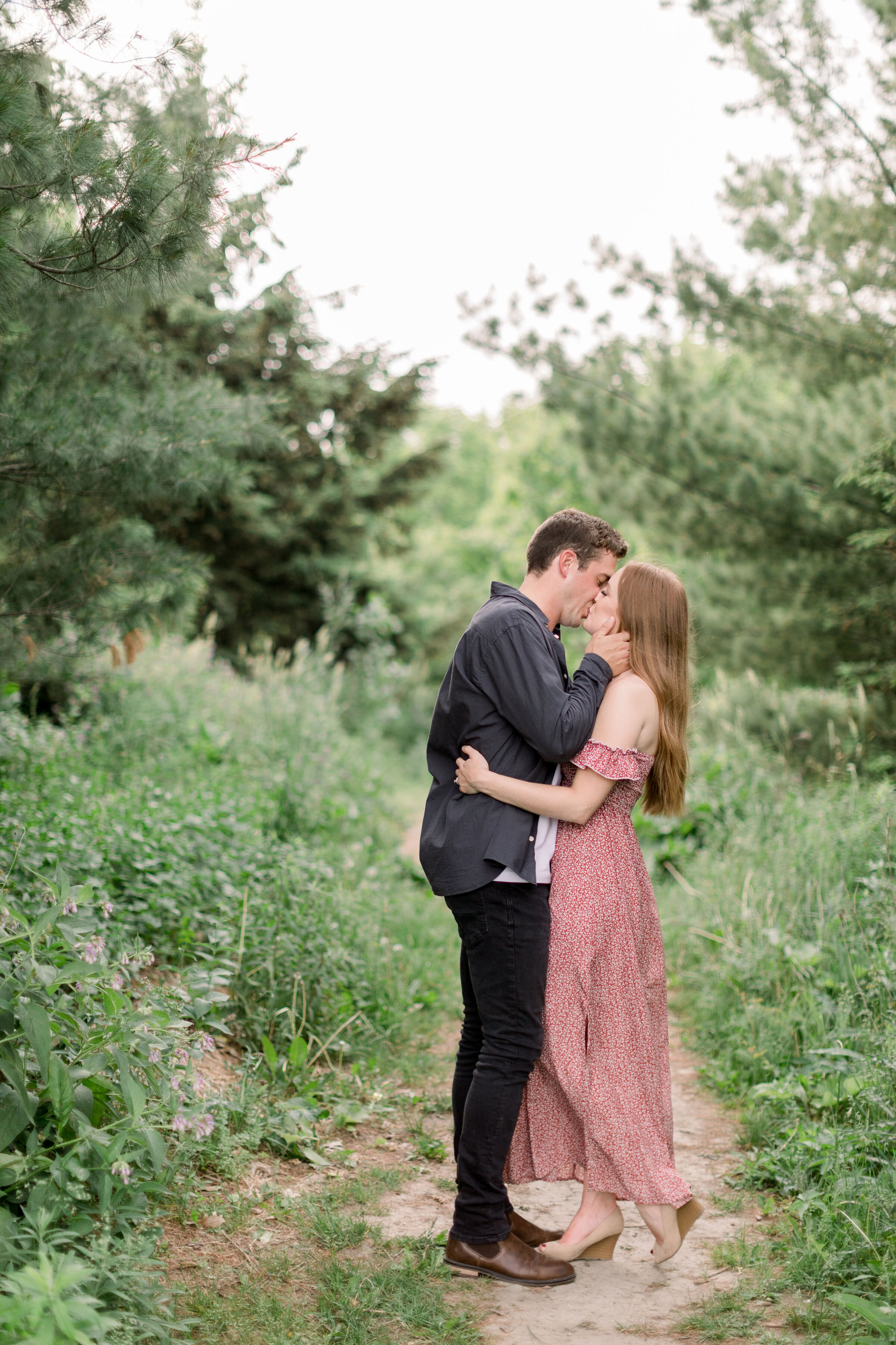  In a shaded unpaved pathway in Arboretum, Ottawa, Chelsea Mason Photography captures this engaged couple romantically kissing. couple kissing woman lifting heel formal engagement outfit inspiration women's off the shoulder maxi dress with nude wedge