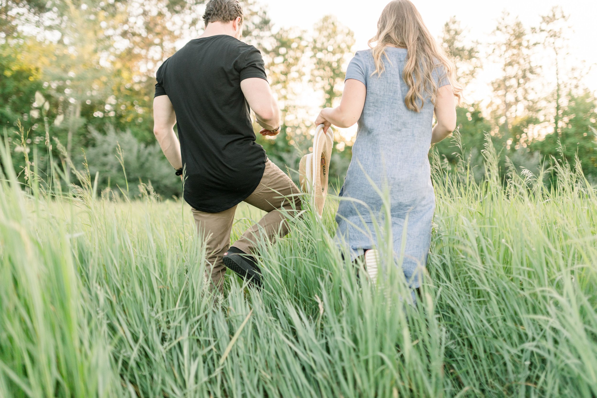  In tall green grass, an engaged couple walks toward the setting sun by Chelsea Mason Photography. outdoor engagement Almonte #chelseamasonphotography #chelseamasonengagements #Ottawaengagments #Almonteengagementphotographer #outdoorengagments 