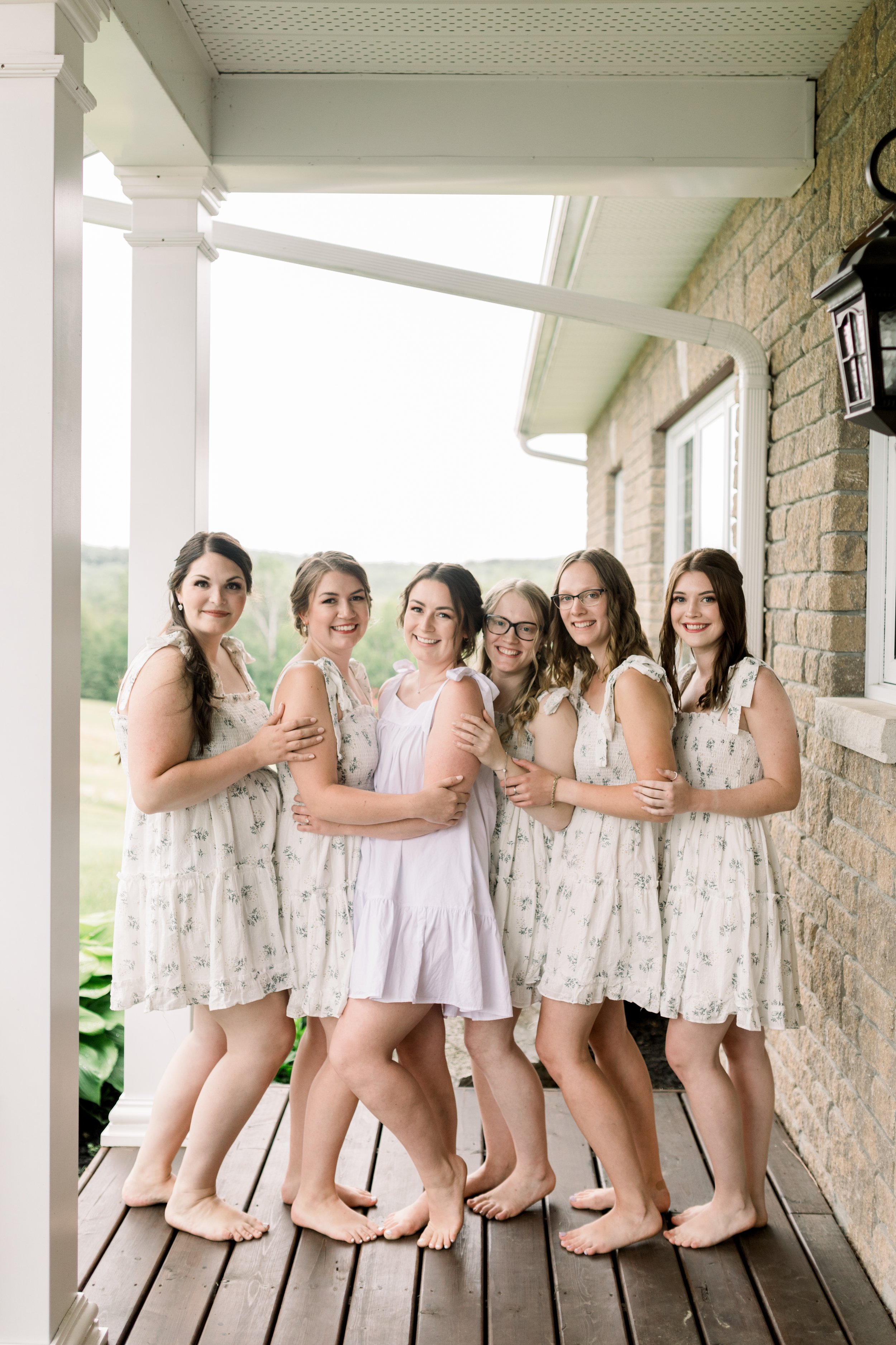  A bride with her bridesmaids on a wooden porch in floral dresses by Chelsea Mason Photography. bridesmaids getting ready porch portrait #chelseamasonphotography #chelseamasonweddings #Ontarioweddings #Boulterweddingphotographer #laceweddinggown 