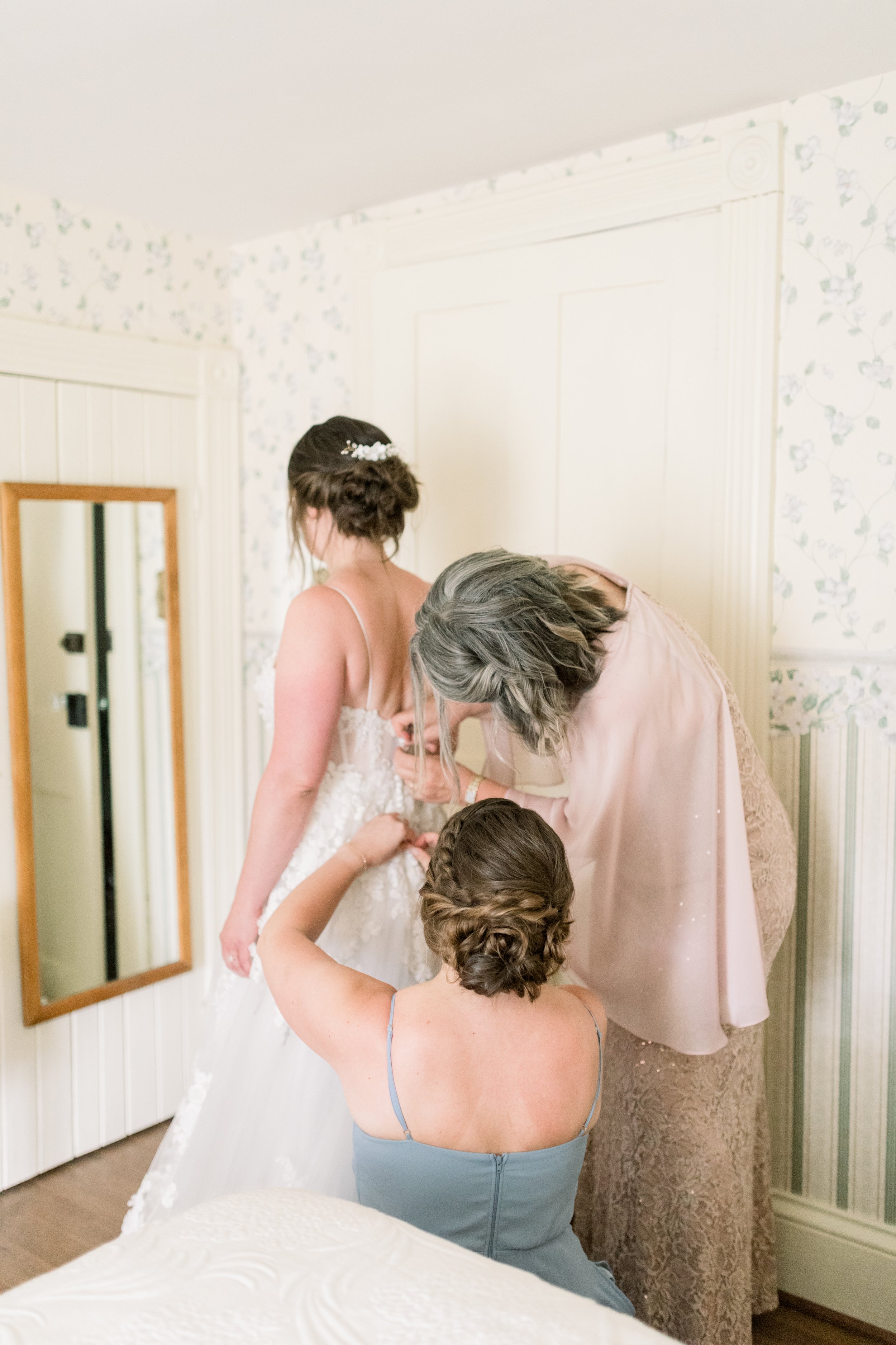  The mother of the bride and sister help the bride put on her wedding gown by Chelsea Mason Photography. mother of the bride wedding gown #chelseamasonphotography #chelseamasonweddings #Ontarioweddings #Boulterweddingphotographer #laceweddinggown 