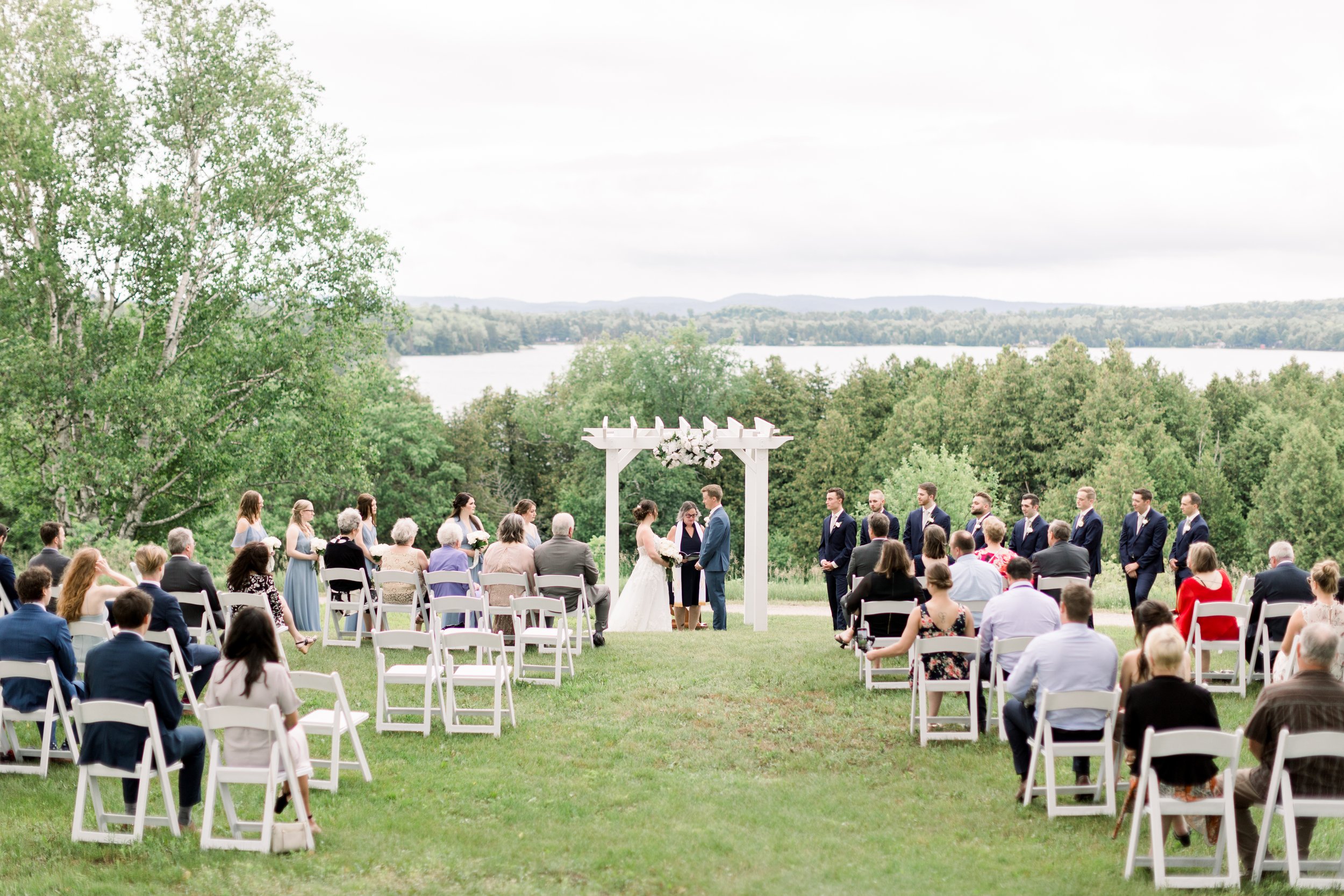  Bride and groom at the altar with wedding guests in the chairs overlooking a lake in Boulter by Chelsea Mason Photography. lake wedding #chelseamasonphotography #chelseamasonweddings #Ontarioweddings #Boulterweddingphotographer #laceweddinggown 