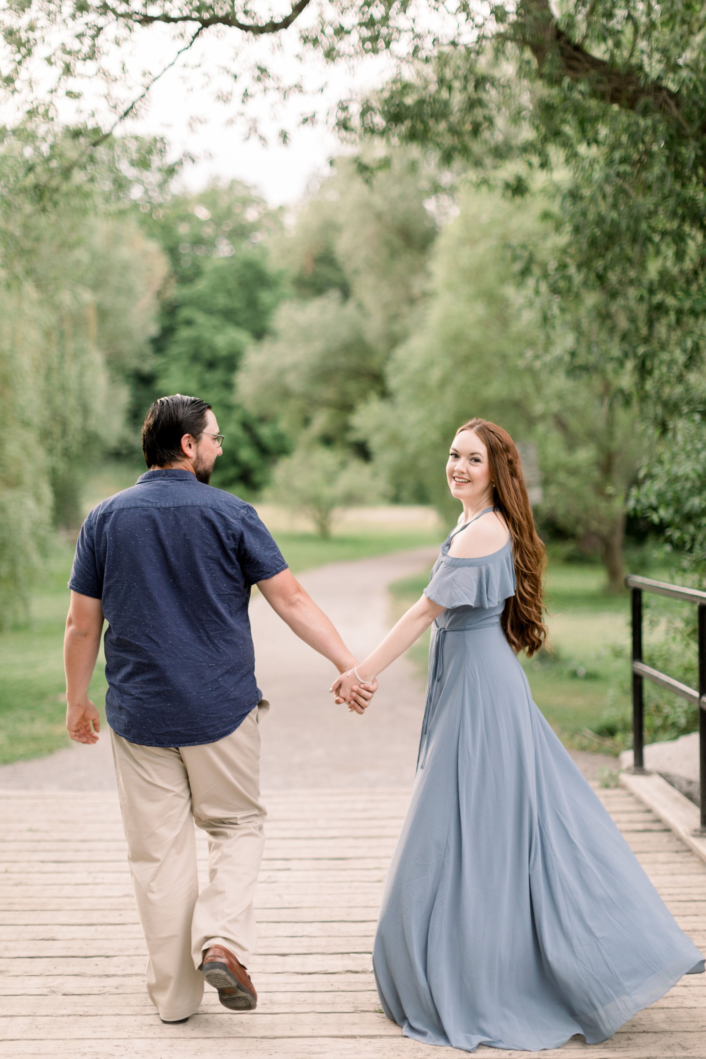  The engaged couple holds hands while walking on a paved path in Ottawa as the woman looks back by Chelsea Mason Photography. paved path #chelseamasonphotography #chelseamasonengagements #Ottawaengagements #DominonArboretum #Ottawaphotographers&nbsp;