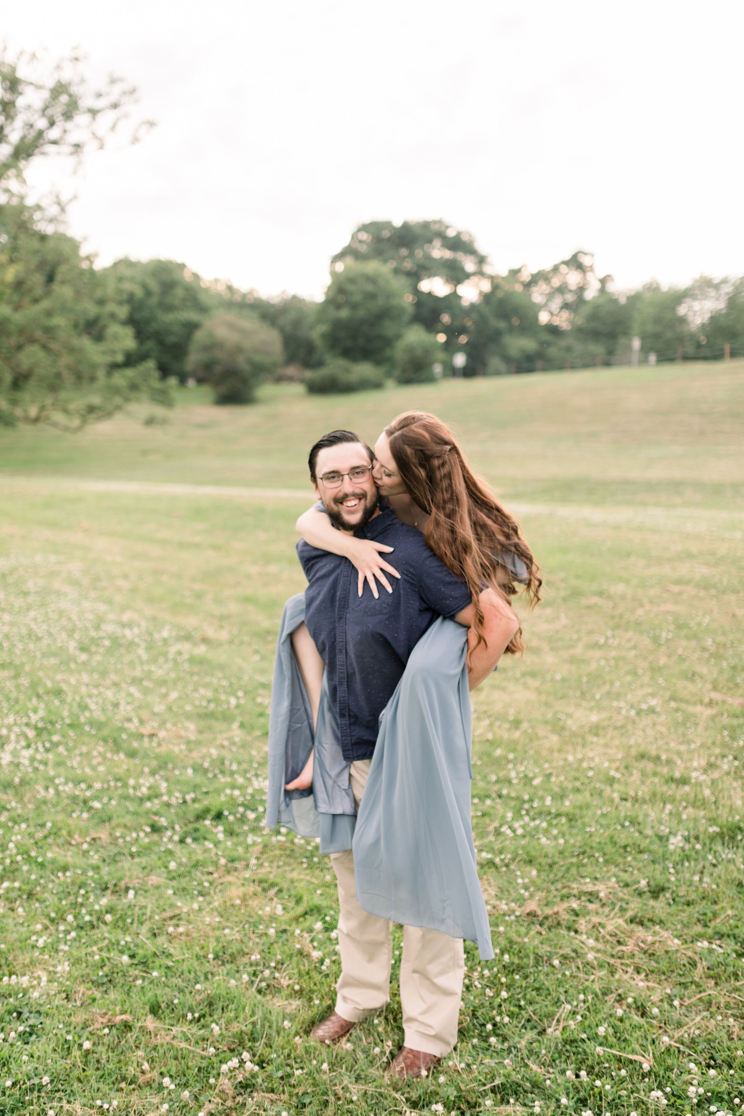  At an Arboretum in Ottawa, a woman hops on her fiances back and kisses his cheek captured by Chelsea Mason Photography. fun engagement poses #chelseamasonphotography #chelseamasonengagements #Ottawaengagements #DominonArboretum #Ottawaphotographers&