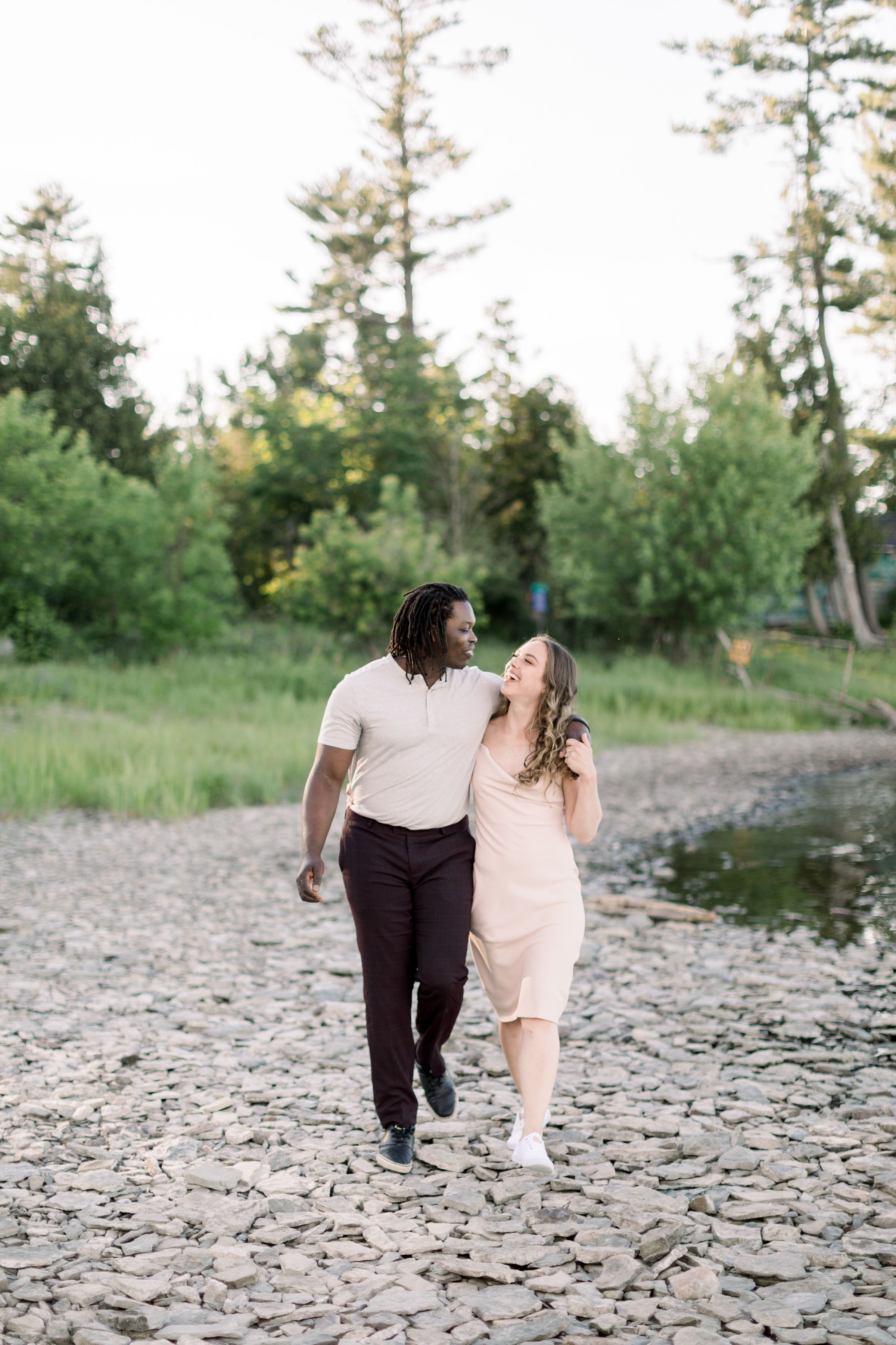  With arms around one another, an engaged couple walks along a stony shore by Chelsea Mason Photography. best friend engagements #Chelseamasonphotography #Chelseamasonengagements #Onatarioengagements #Pinhey'sPoint #Ontarioweddingphotographers 