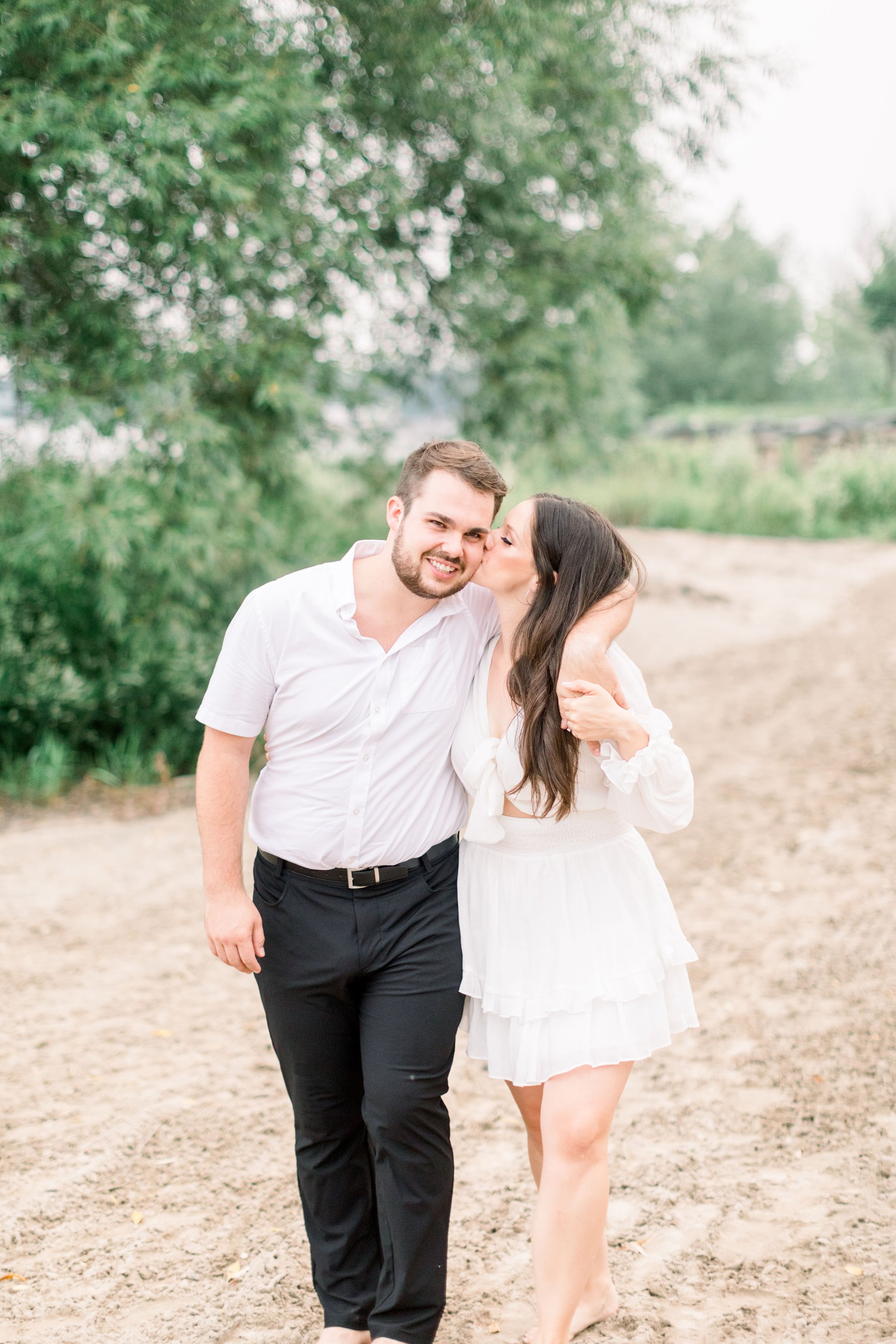  Walking with his arm around his fiance a man receives a kiss on the cheek by Chelsea Mason Photography. woman kiss man #Kingstonengagement #GrassCreekParkPortraits #Ontarioengagementphotographers #Chelseamasonphotography #Chelseamasonengagements 
