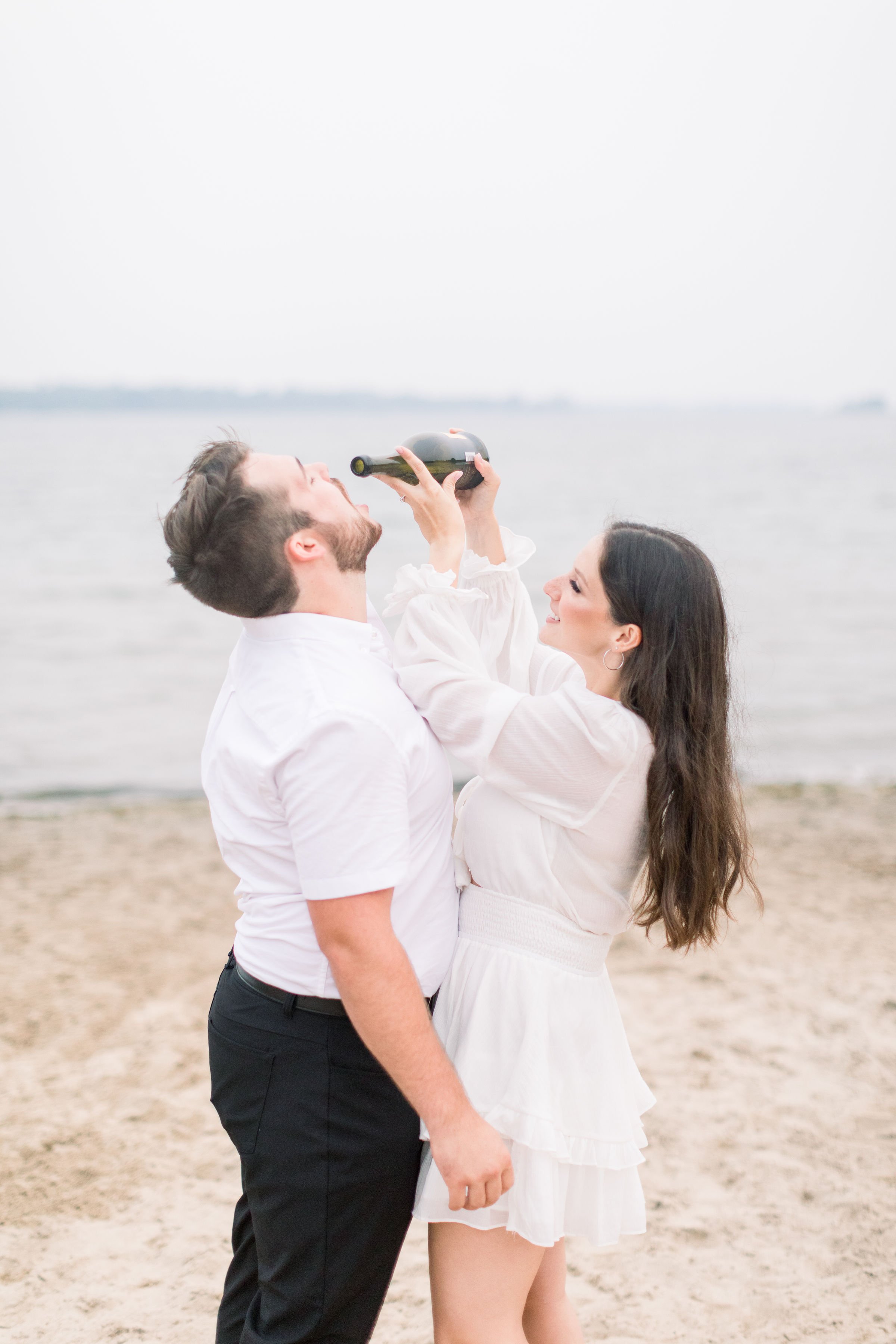  On a lakeshore, a woman pours a bottle of champagne into her fiance’s mouth by Chelsea Mason Photography. white dress summer #Kingstonengagement #GrassCreekParkPortraits #Ontarioengagementphotographer #Chelseamasonphotography #Chelseamasonengagement