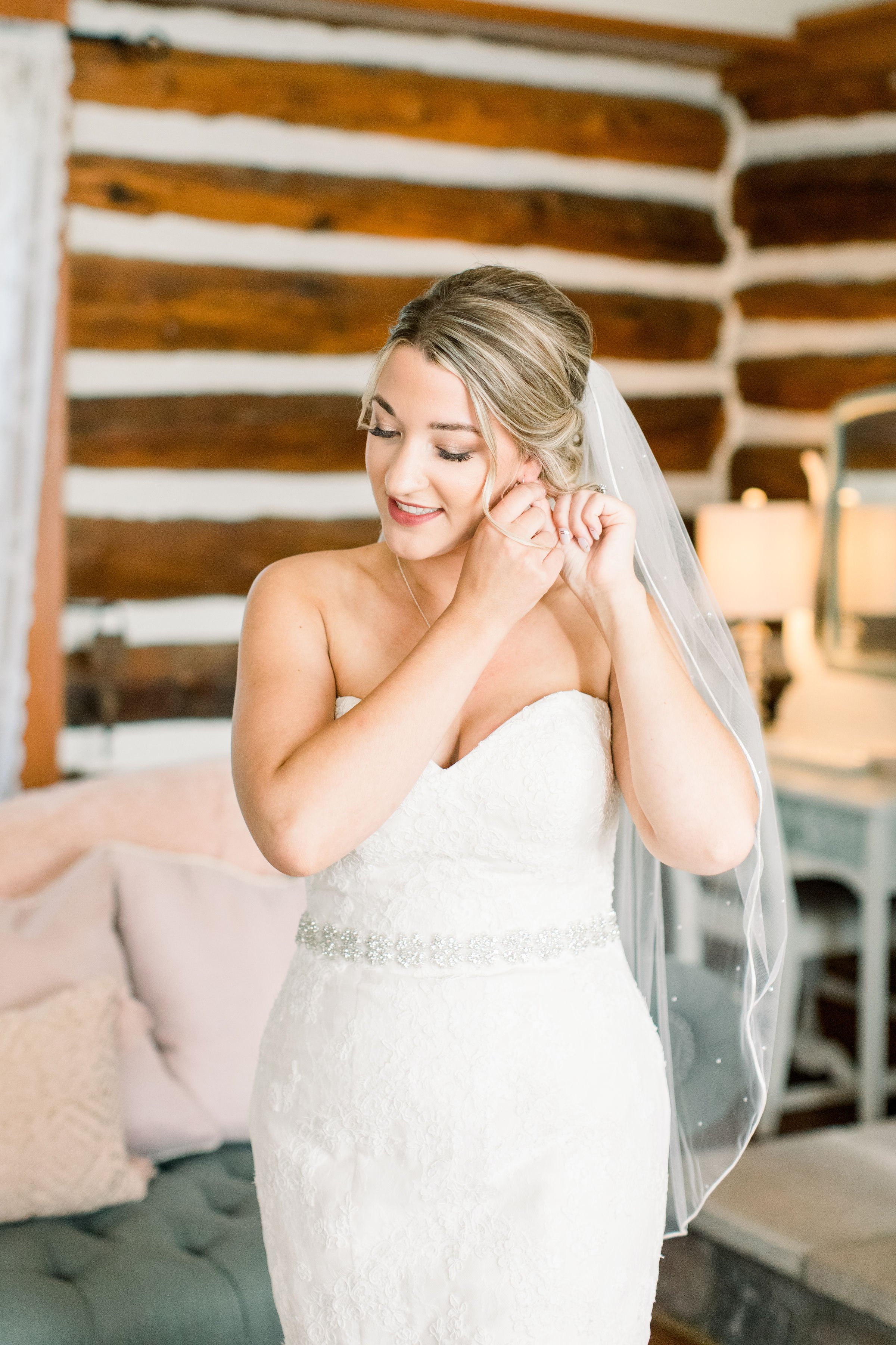  A bride wearing a strapless wedding gown puts on her earring in a cabin by Chelsea Mason Photography. form-fitting wedding dress #StonefieldEstates #Ontarioweddings #Chelseamasonphotography #Chelseamasonengagements #Ontarioweddingphotographers 