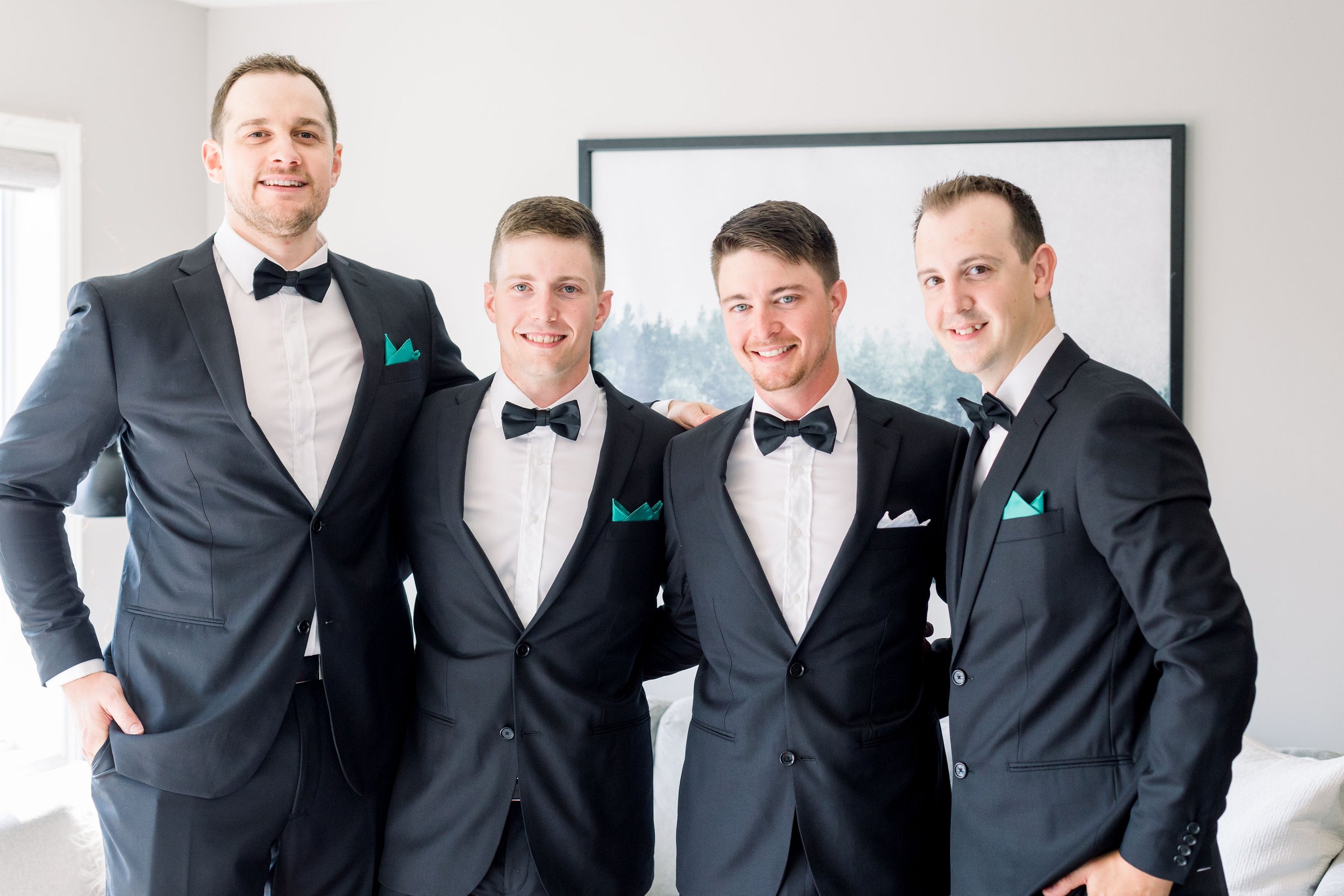  A groom smiles with his three groomsmen all wearing black suits and bowties by Chelsea Mason Photography. men classic wed style #ChelseaMasonPhotography #ChelseaMasonWeddings #PrinceEdwardsCountyWeddings #SandbanksWeddings #ONweddingphotographer 