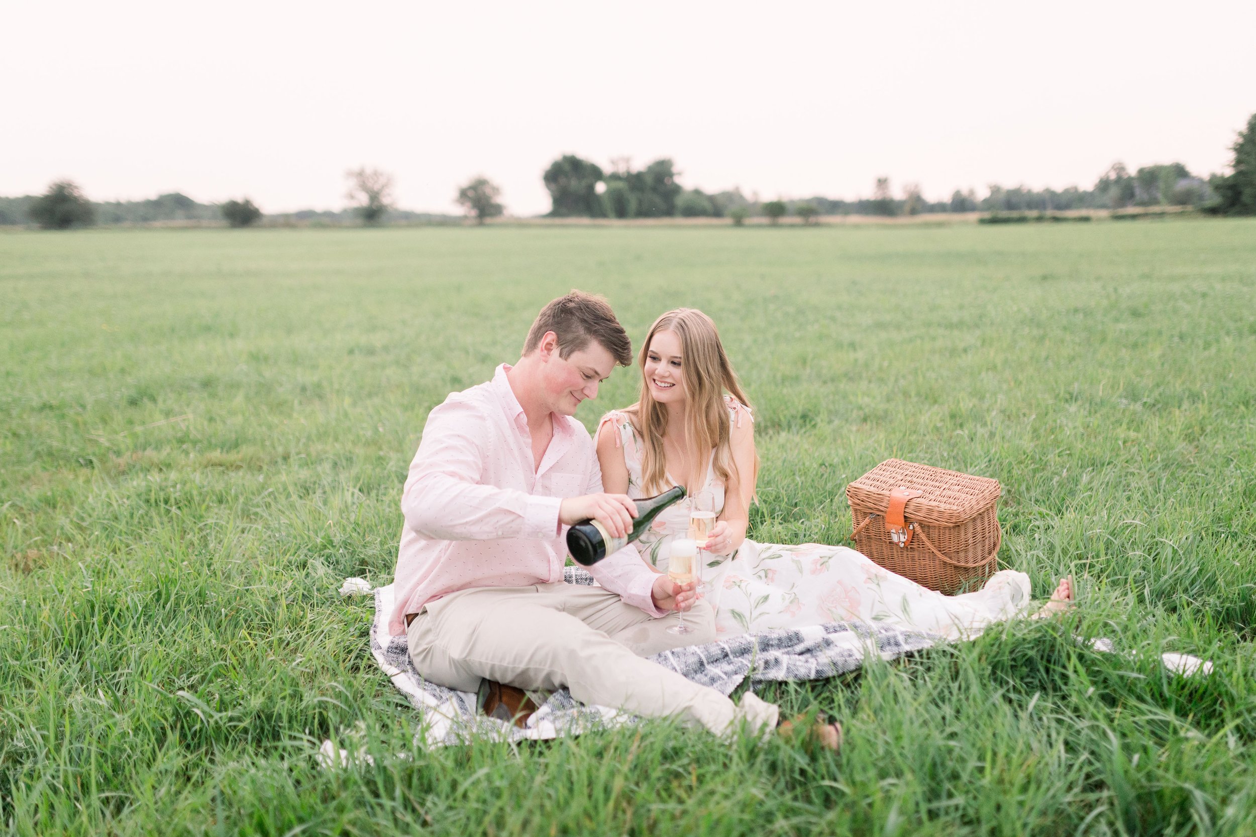  Fiances celebrate with champagne sitting on a picnic blanket in a grass field by Chelsea Mason photography. Picnic wedding celebration #ChelseaMasonPhotography #ChelseaMasonEngagements #PinheysPointEngagements #OttawaEngagementPhotography 