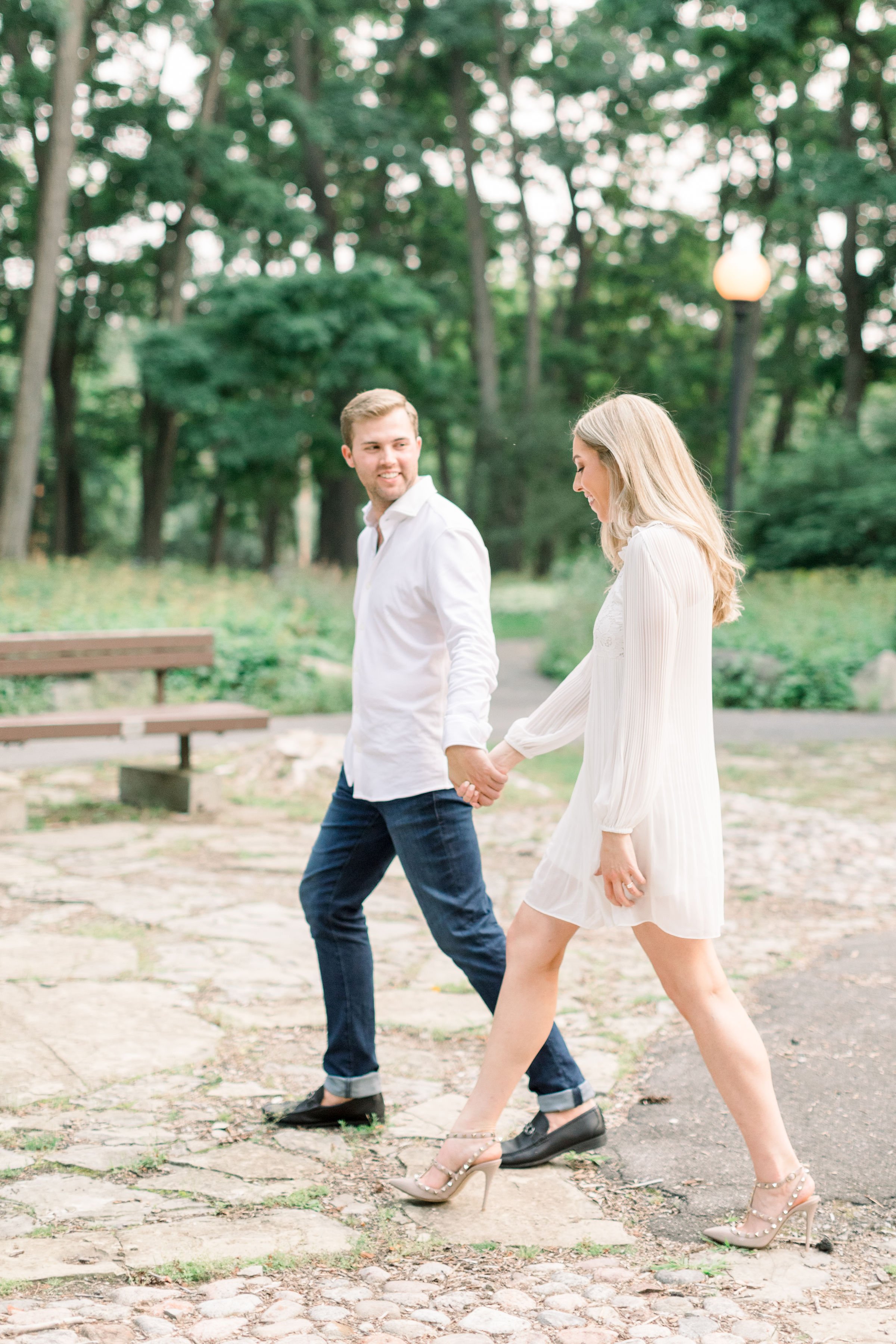  Walking hand in hand fiances smile at one another by Chelsea Mason Photography an engagement photographer. stone path white dress #Ottawaengagements #Ottawaweddingphotographers #engagementwithdogs #ChelseaMasonPhotography #ChelseaMasonEngagements  