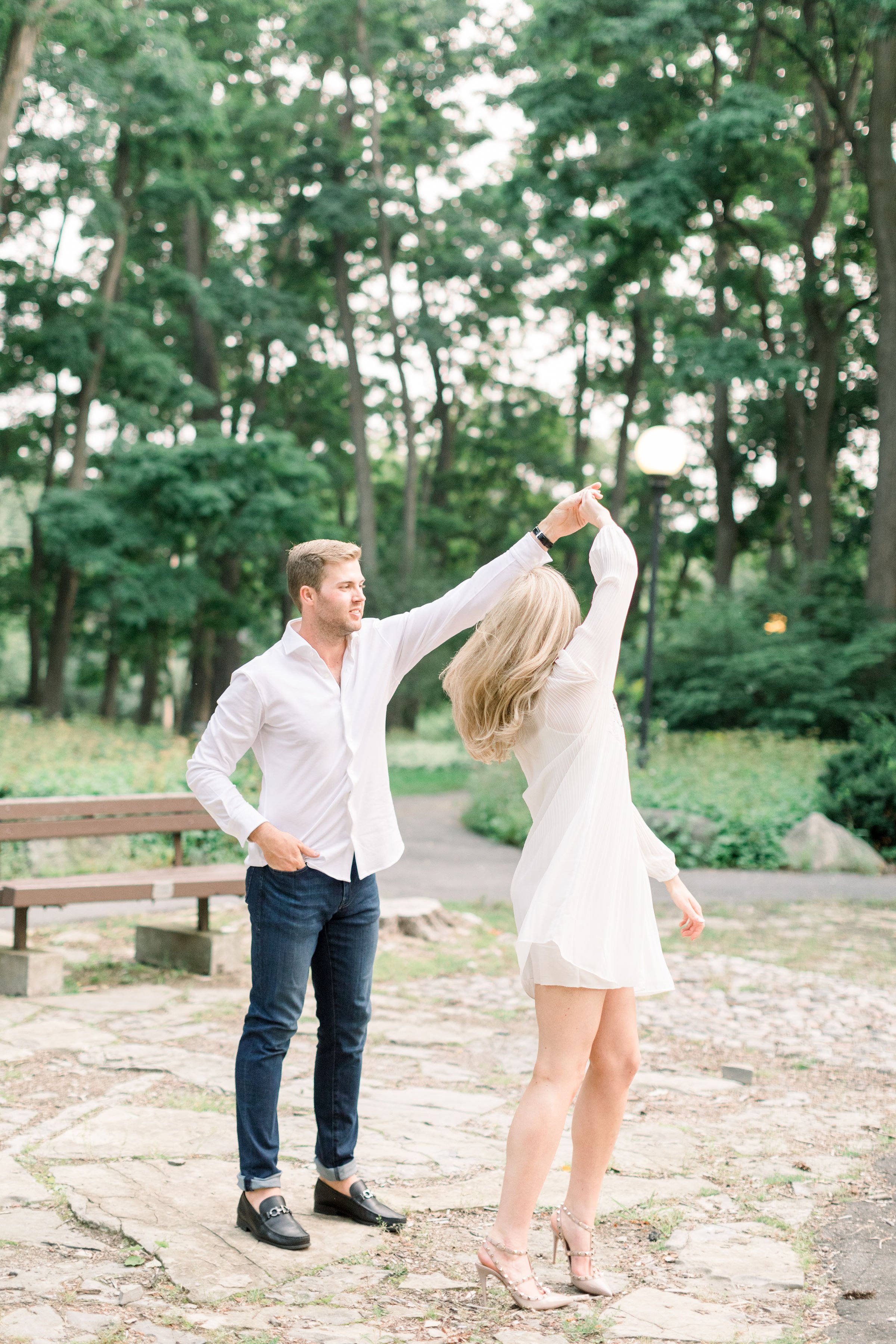  A man twirls his bride-to-be next to a bench captured by Chelsea Mason Photography. dancing with a boy in white outfits #Ottawaengagements #Ottawaweddingphotographers #engagementwithdogs #ChelseaMasonPhotography #ChelseaMasonEngagements  