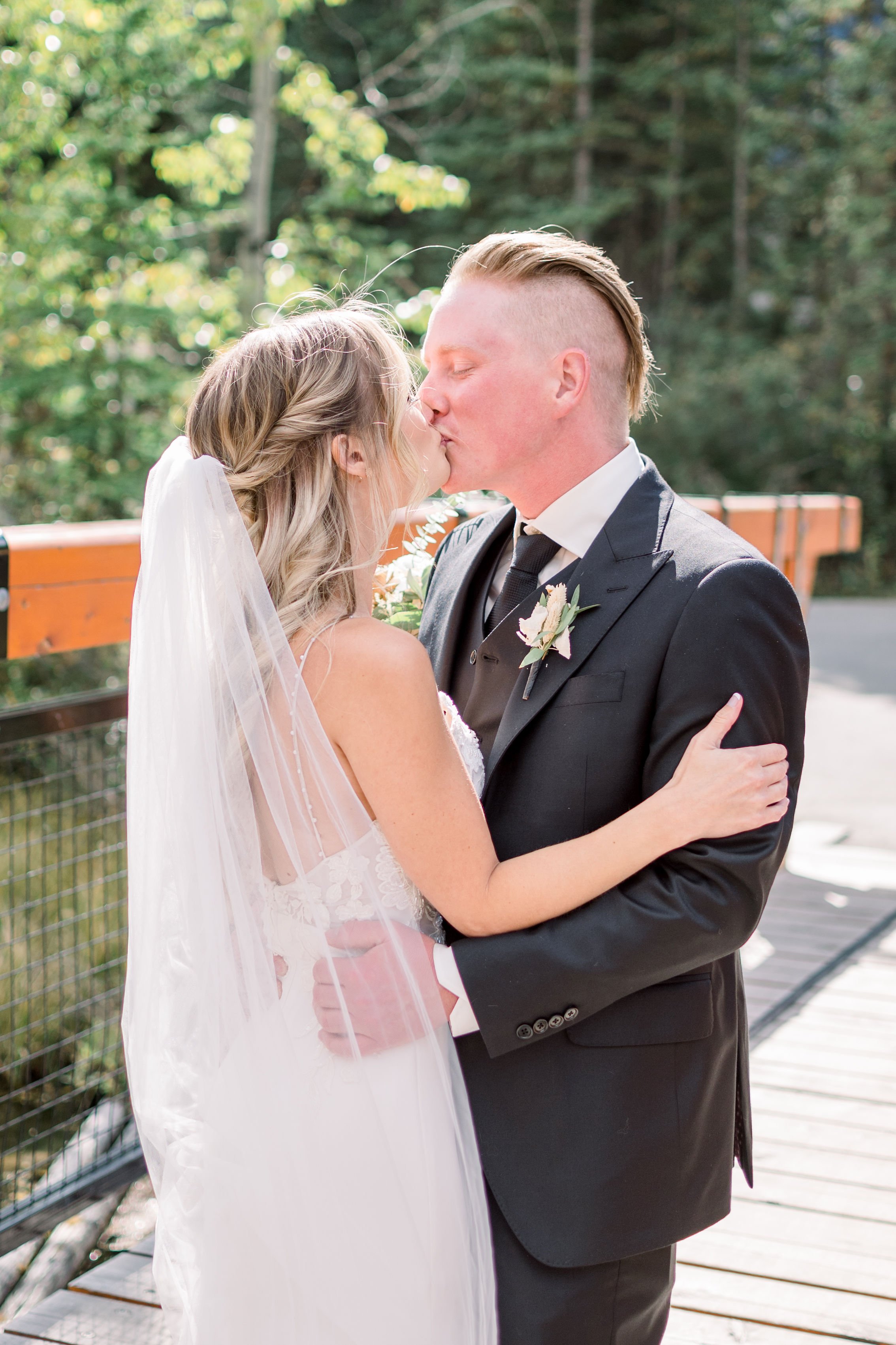  Newlyweds kiss on a summer day in the mountains of Banff by Chelsea Mason Photography. black suit and tie country wedding #Albertaweddings #shesaidyes #Albertaweddingphotographers #SilvertipGolfCourse #ChelseaMasonPhotography #ChelseaMasonWeddings  