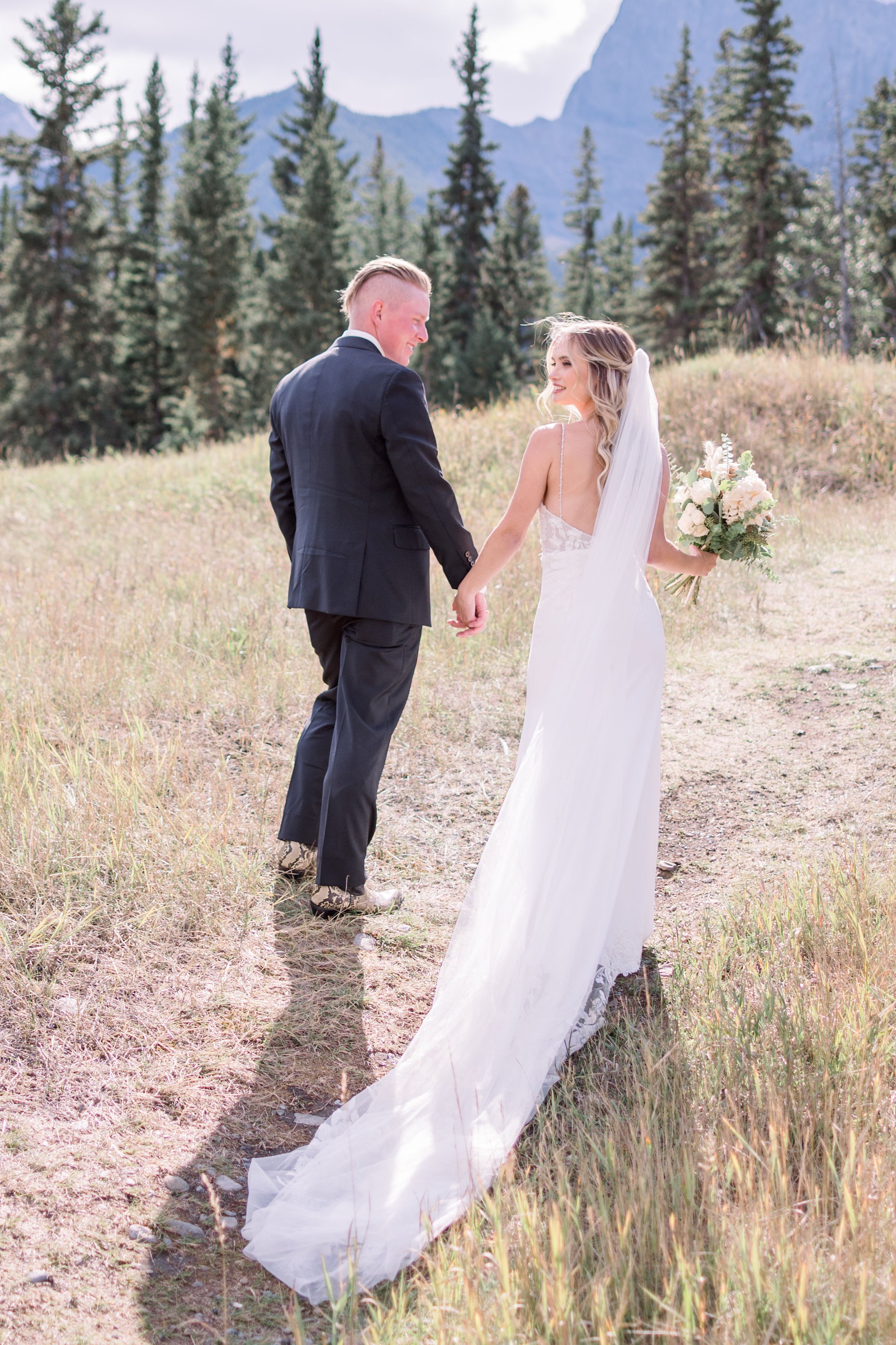  A bride and groom walk up a mountain pathway of yellow grass by Chelsea Mason Photography. mountain bridals Silvertip Golf #Albertaweddings #shesaidyes #Albertaweddingphotographers #SilvertipGolfCourse #ChelseaMasonPhotography #ChelseaMasonWeddings 