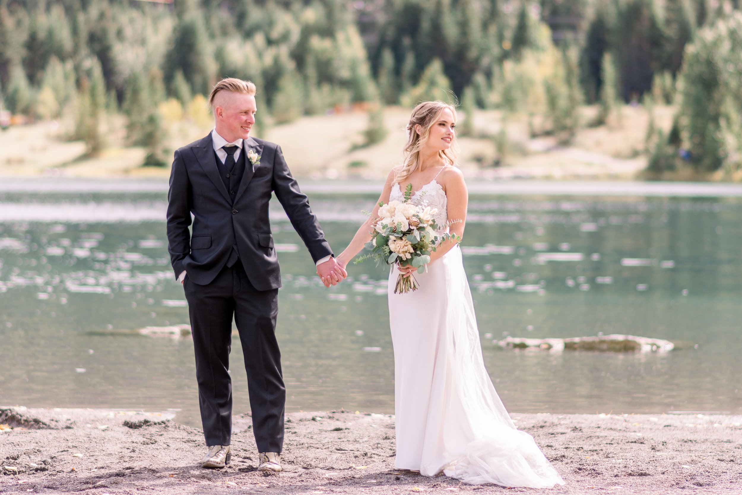  In Alberta, Canada a bride and groom hold hands next to a mountain lake by Chelsea Mason Photography. country wed outdoor #Albertaweddings #shesaidyes #Albertaweddingphotographers #SilvertipGolfCourse #ChelseaMasonPhotography #ChelseaMasonWeddings  