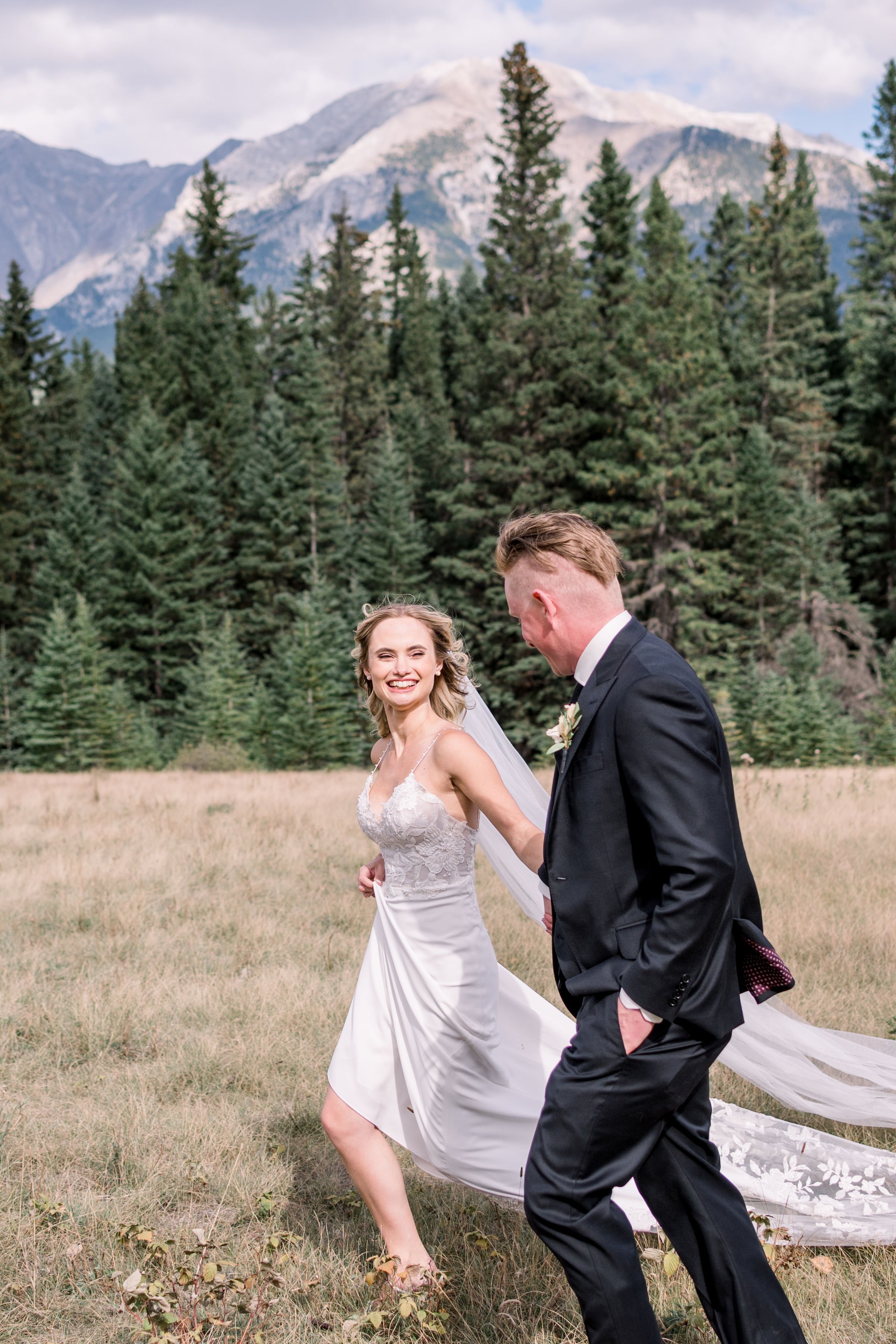  With pine trees behind them, a bride and groom run through a field by Chelsea Mason Photography. Banff weddings #Albertaweddings #shesaidyes #Albertaweddingphotographers #SilvertipGolfCourse #ChelseaMasonPhotography #ChelseaMasonWeddings  