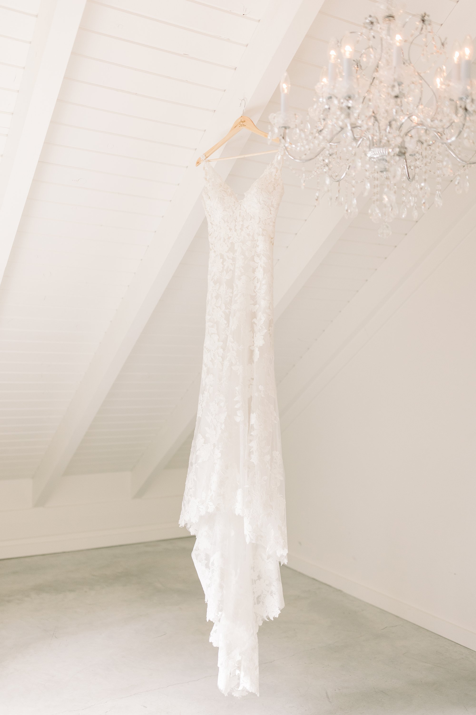  A vintage lace bridal gown hangs in a white attic next to a chandelier by Chelsea Mason Photography. vintage bridal gown #ChelseaMasonPhotography #ChelseaMasonWeddings #WakefieldWeddings #LeBelvedere #Wakefieldweddingphotographers 