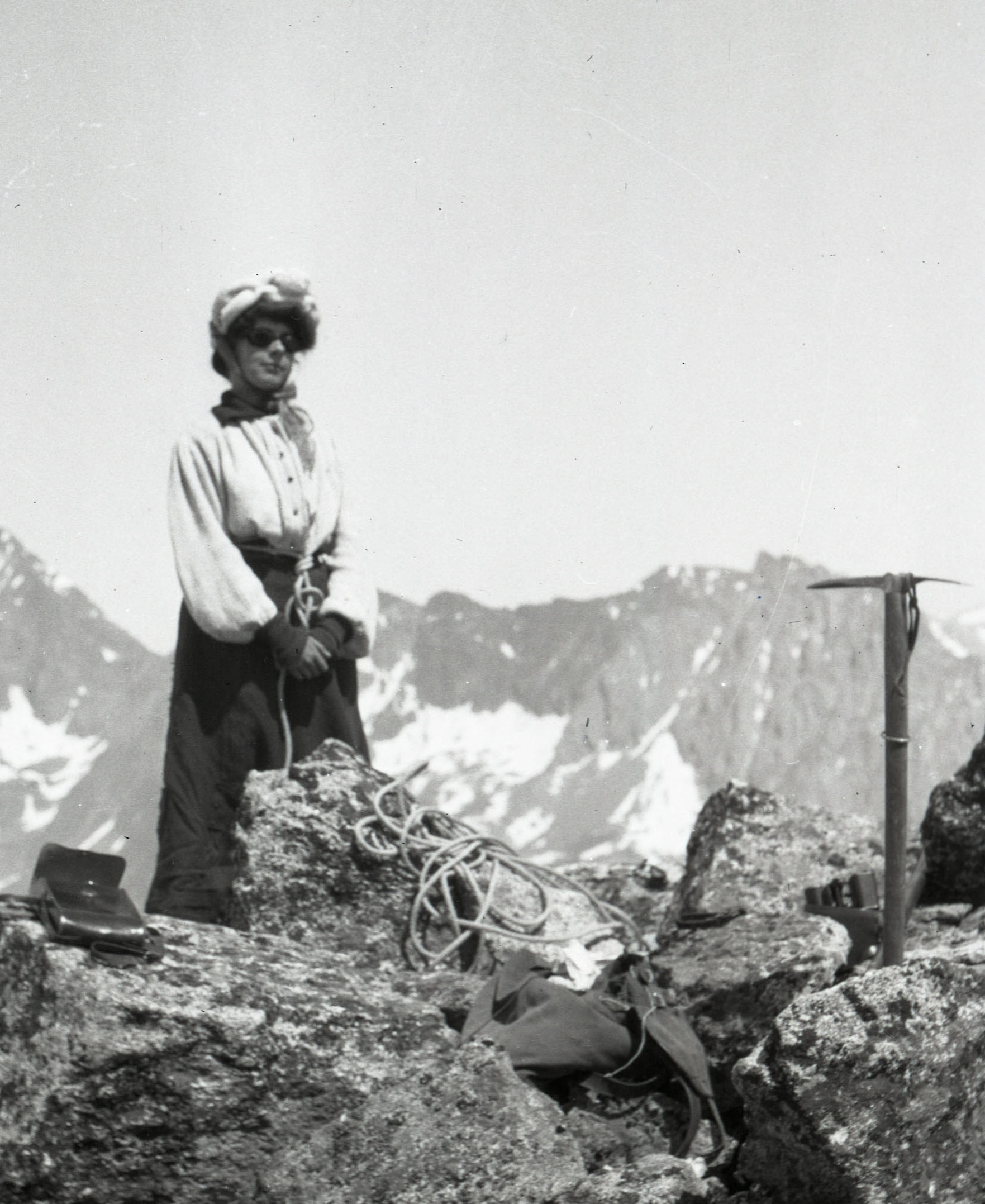 Photo credit: American Alpine Club Library. From our Andrew J. Gilmour collection, taken in the Alps around 1910. A woman in a long coat and glacier goggles stands in front of a narrow rock prominence. She is holding an ice axe and is part of a rope team, roped to other mountaineers.