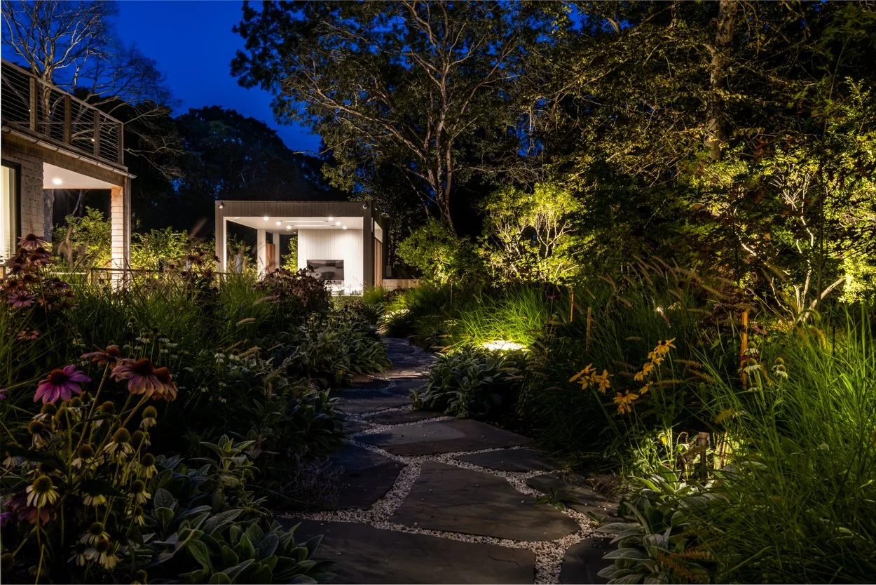 Garden lighting composition at Amagansett estate — warm uplighting on mature trees and garden beds at night