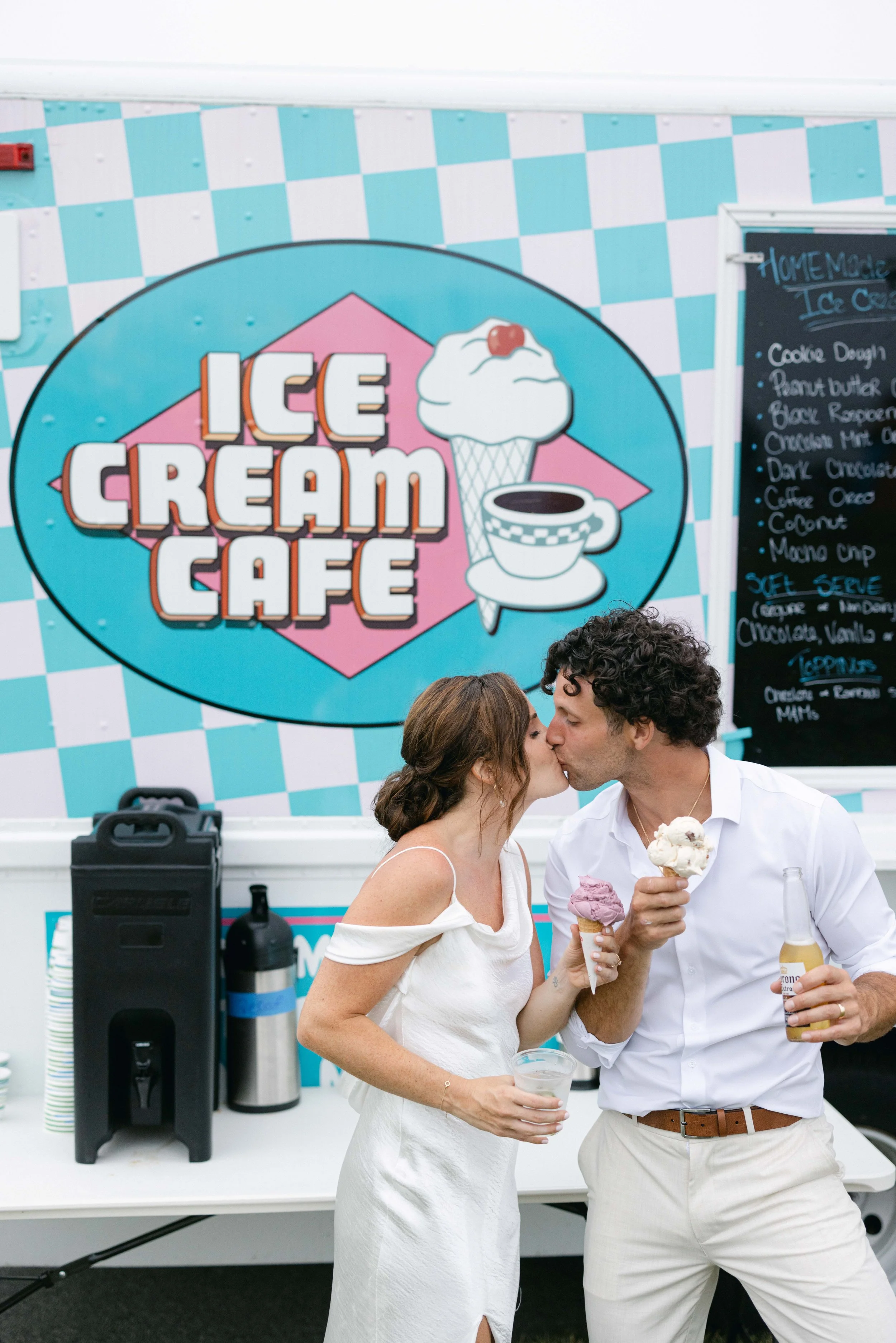 A couple kisses while holding ice cream cones at an ice cream cafe, with a large colorful sign behind them that reads 'Ice Cream Cafe'.