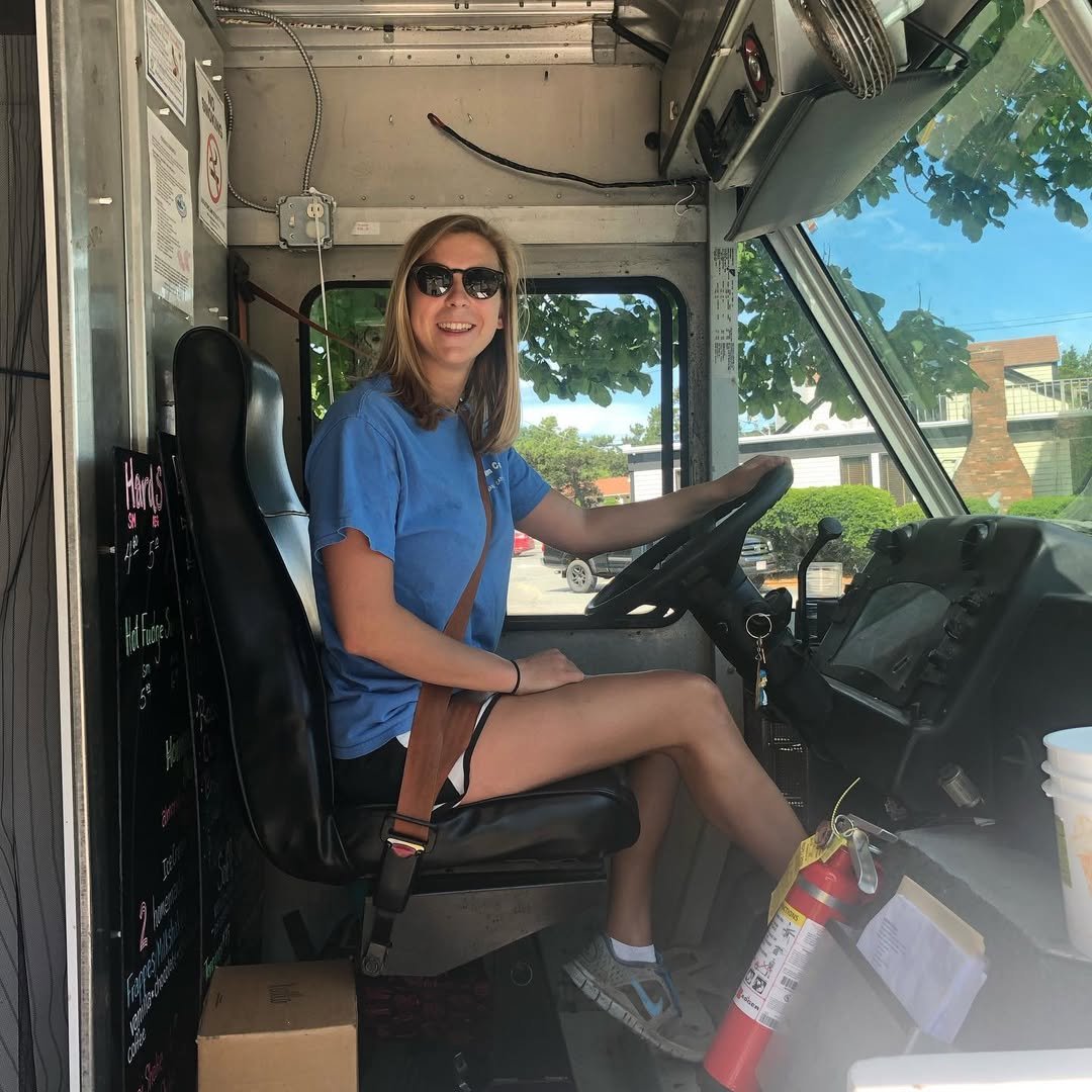 A woman wearing sunglasses, a blue shirt, and shorts, sitting in the driver’s seat of a food truck, smiling at the camera.