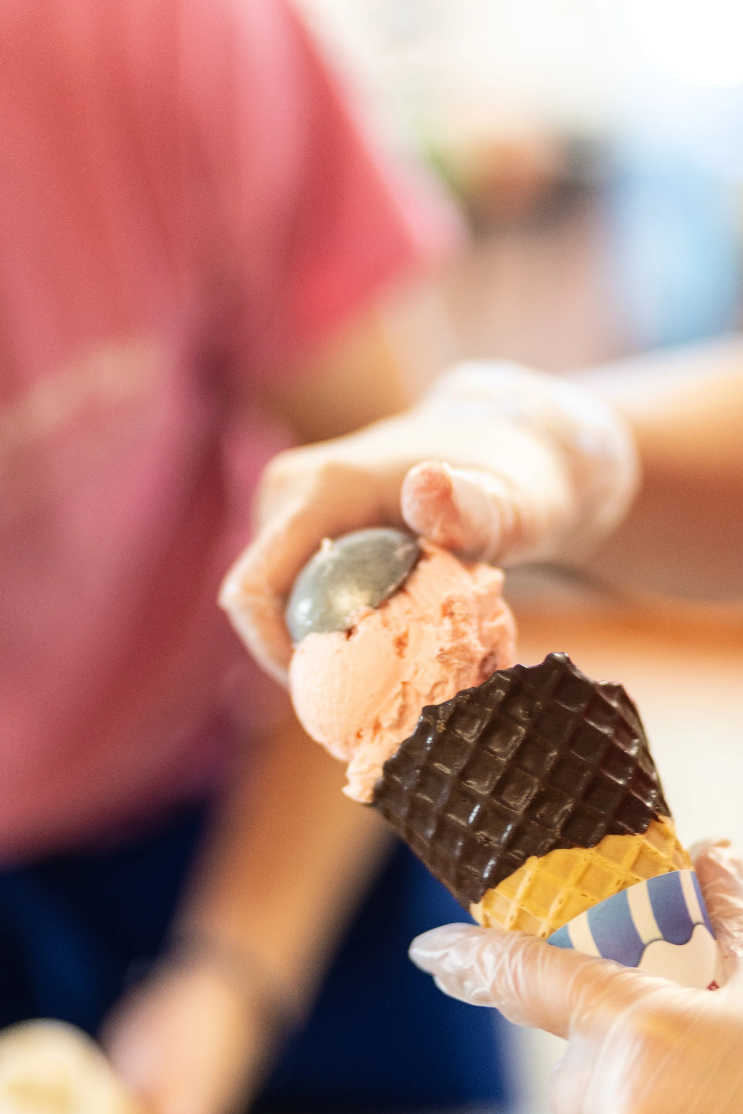 Close-up of a person holding a small ice cream cone with two scoops of peach-colored ice cream, one having a small blue object on top, and dark chocolate topping on the cone.