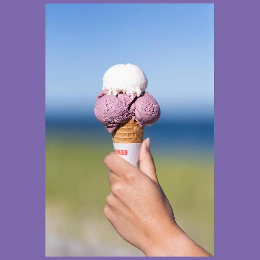 Hand holding a three-scoop ice cream cone with purple and white ice cream, against a blurred outdoor background.