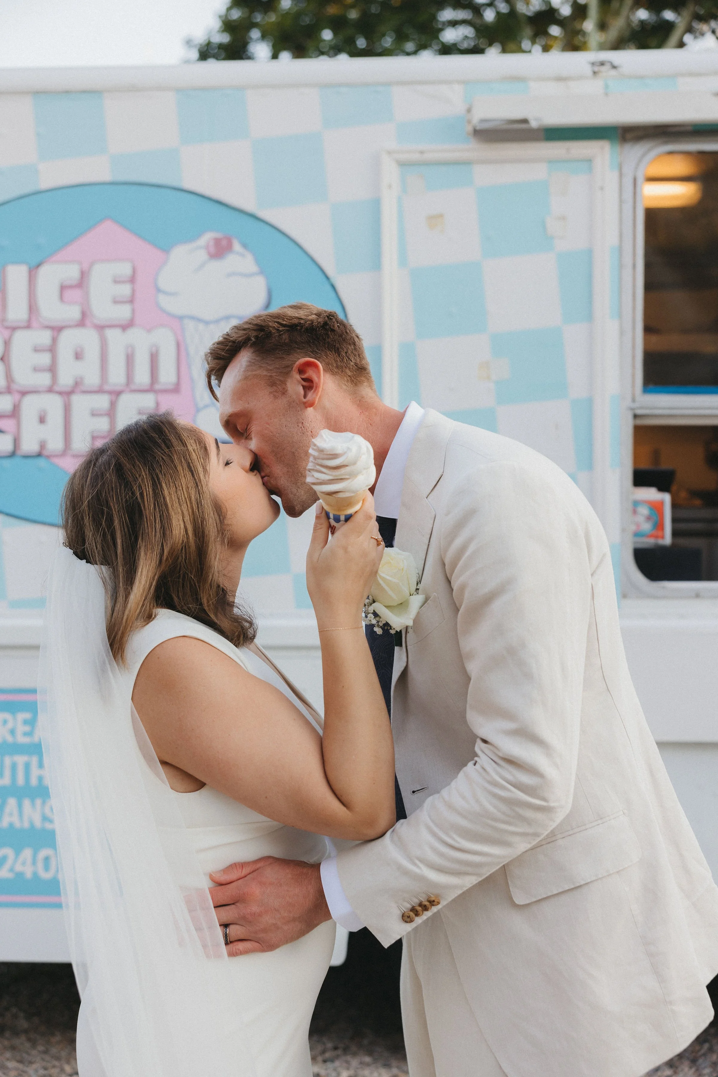 A bride and groom sharing a kiss while holding an ice cream cone in front of an ice cream truck.