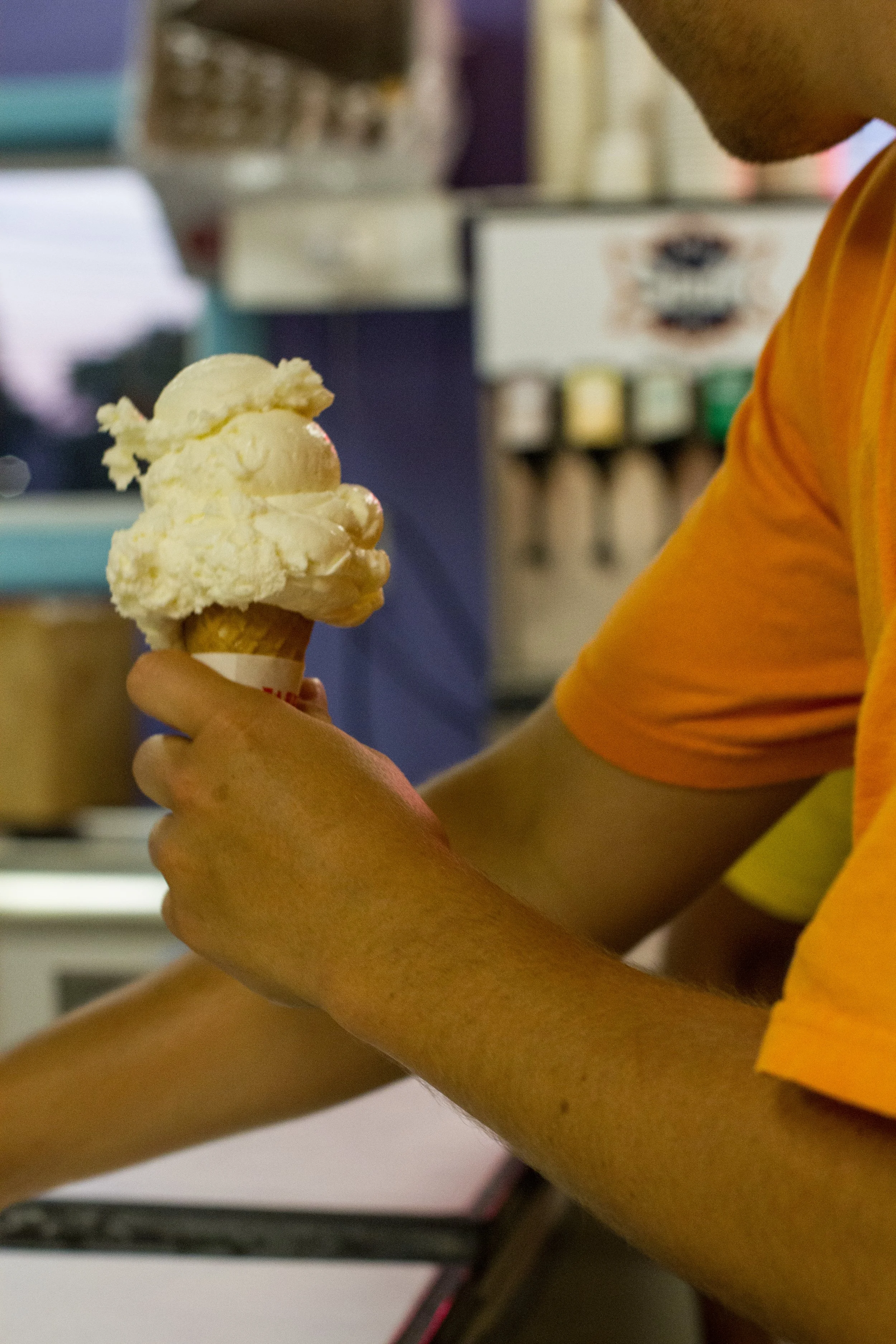 Person in an orange shirt holding a single scoop of vanilla ice cream in a cone inside an ice cream shop.