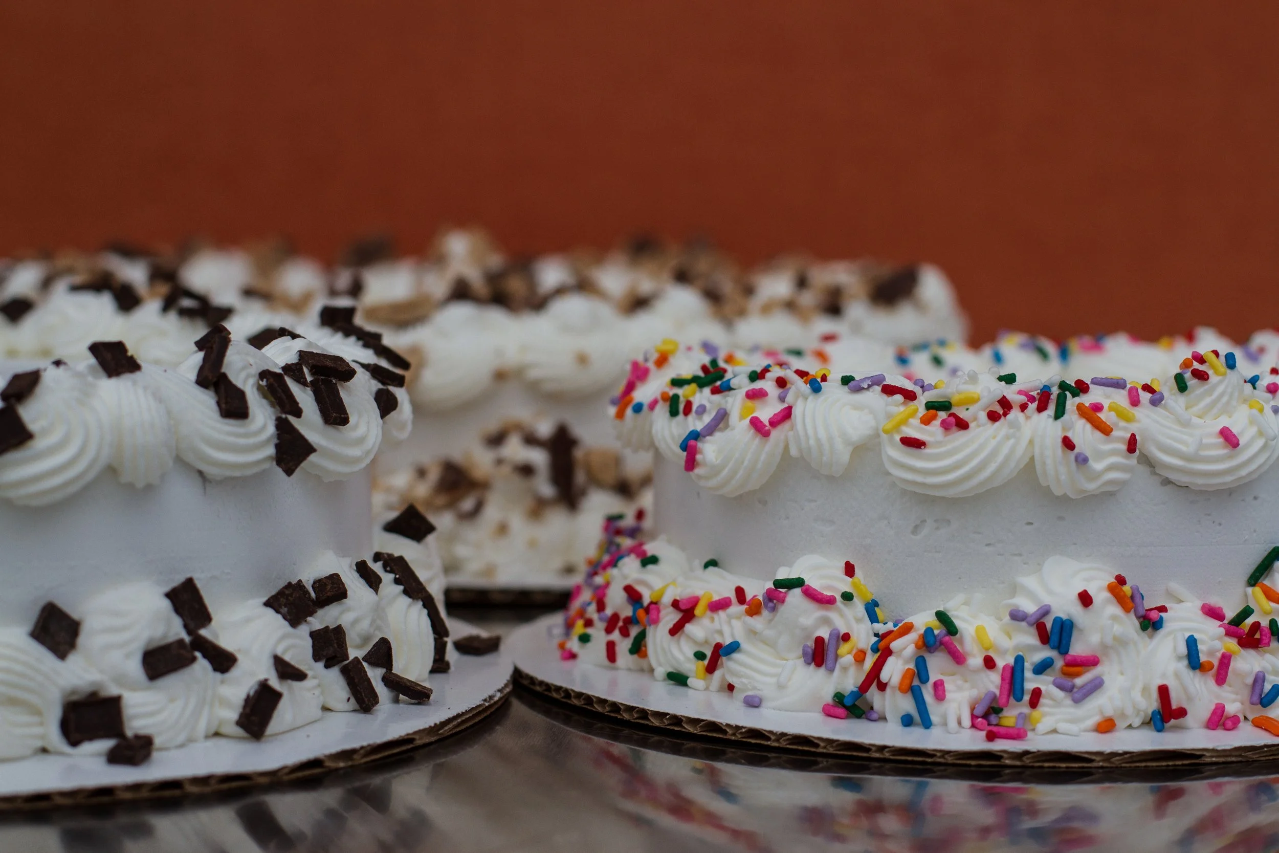 Three decorated birthday cakes with white frosting and colorful sprinkles or chocolate shavings on top.