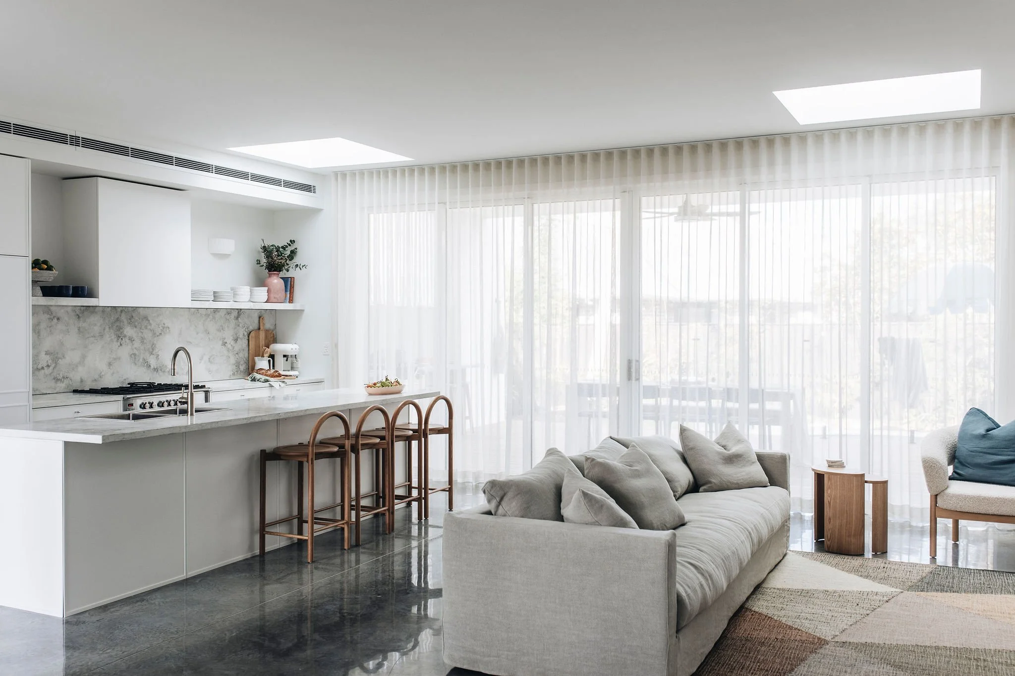 Bright, modern living room and open kitchen with white cabinets, a marble island with wooden bar stools, a gray sofa, and sliding glass doors with sheer curtains.