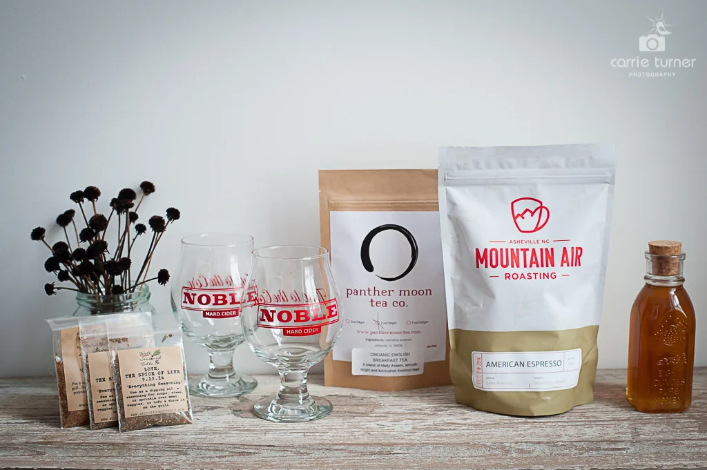 A collection of tasty food and drink favorites: spice mix samples from The Well Seasoned Table, two Belgian-style cider glasses from Noble Cider, a bag of Organic English Breakfast Tea from the Panther Moon Tea Company, a bag of American Espresso from Mountain Air Roasting Company, and a bottle of Basswood/Sourwood honey from Wild Mountain Apiaries, carried by the Asheville Bee Charmer. 