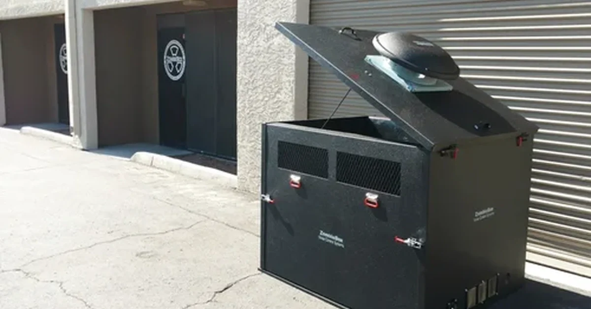 Black metal generator enclosure sits outside by a roll-up door, lid open with a round roof fan in bright sunlight.