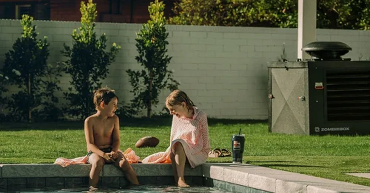 A poolside area with two children sitting at the edge with their feet in the water beside a lawn and equipment box.