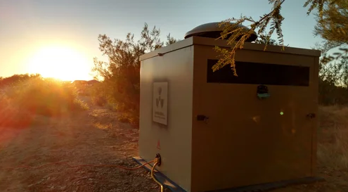 A tan generator enclosure sits in a dry desert area with cables on the ground and low sunlight behind trees.