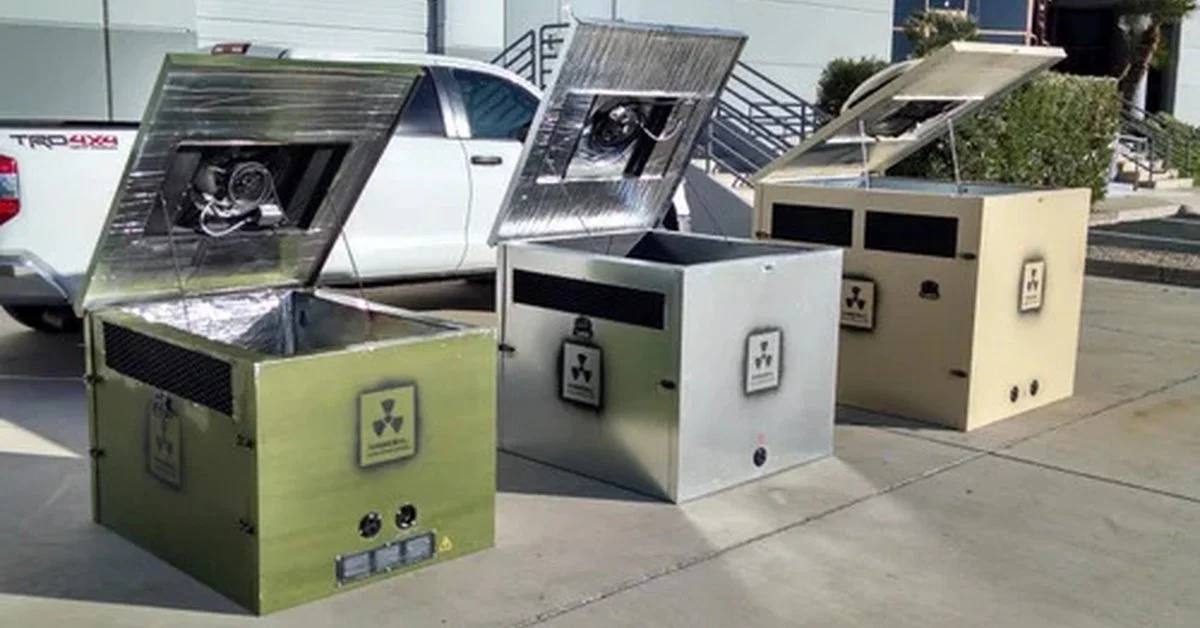 Three metal equipment enclosures with raised lids are lined on pavement beside a white pickup truck and shrubs.