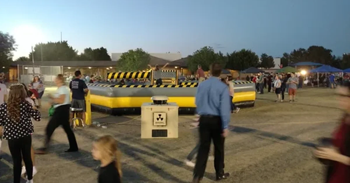 People walk by an outdoor event near a yellow-and-black inflatable arena, with a generator enclosure set up nearby.