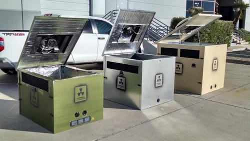 Three metal generator enclosures with raised lids and visible fans standing on pavement beside a white pickup truck.