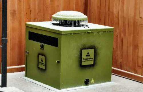 An olive green generator enclosure stands on a concrete pad by a wooden fence, with a round capped top edged in ice.