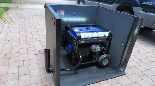 A portable generator sits inside an open acoustic panel on a paved driveway beside a parked gray truck.