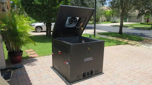 A large black outdoor generator silencer with an open lid standing on a brick patio beside grass, trees, and potted plants.