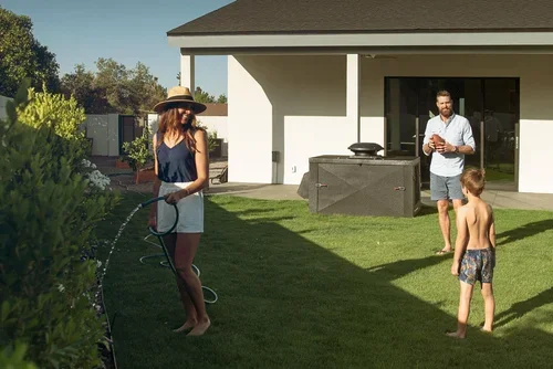 A woman watering shrubs as a man and child stand on a lawn near a black outdoor equipment box by a house.