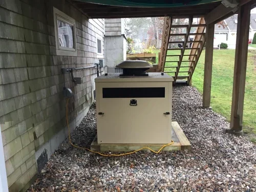 A cream generator unit sitting under a raised deck beside a shingled building, gravel, grass, and wooden stairs.