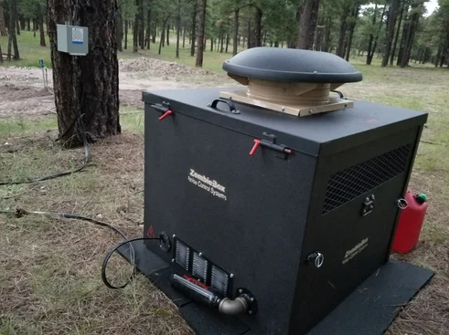 A tan generator enclosure sits in a dry desert area with cables on the ground and low sunlight behind trees.