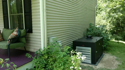 An outdoor generator with an enclosure sits beside a house near a porch, partially hidden by leafy shrubs in summer.